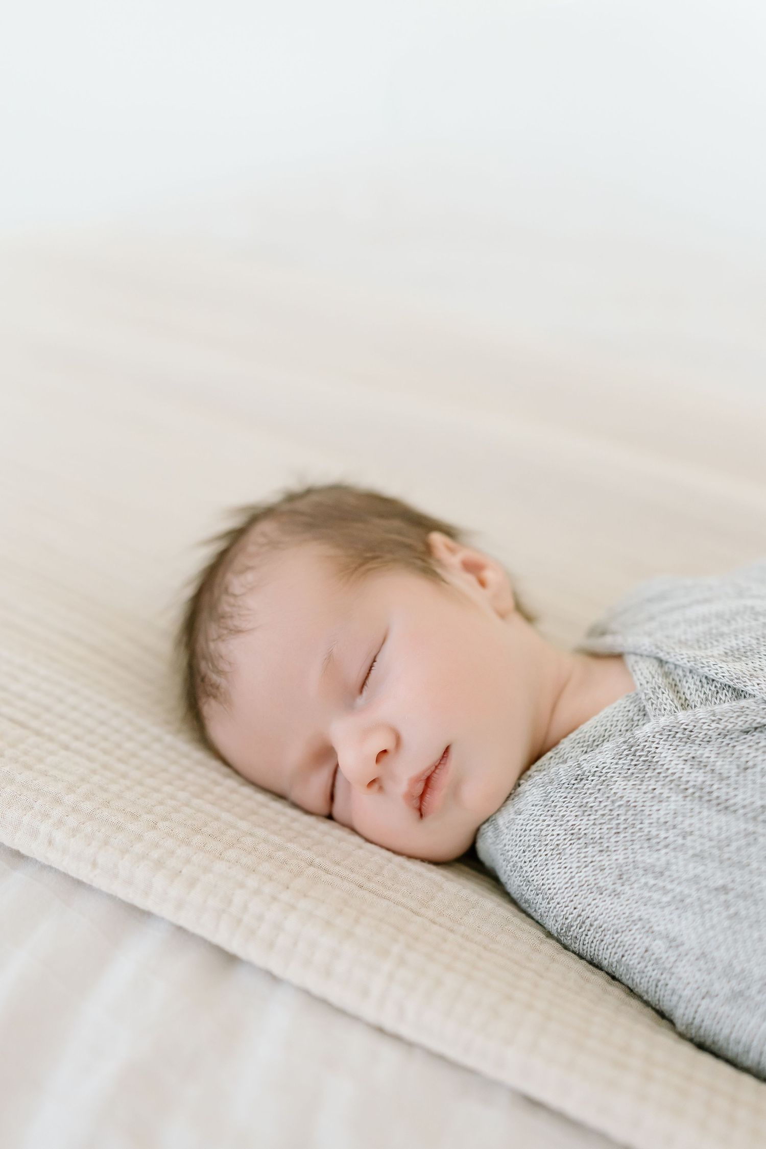 A sleeping baby wearing a gray sweater rests peacefully on a textured cream-colored blanket.