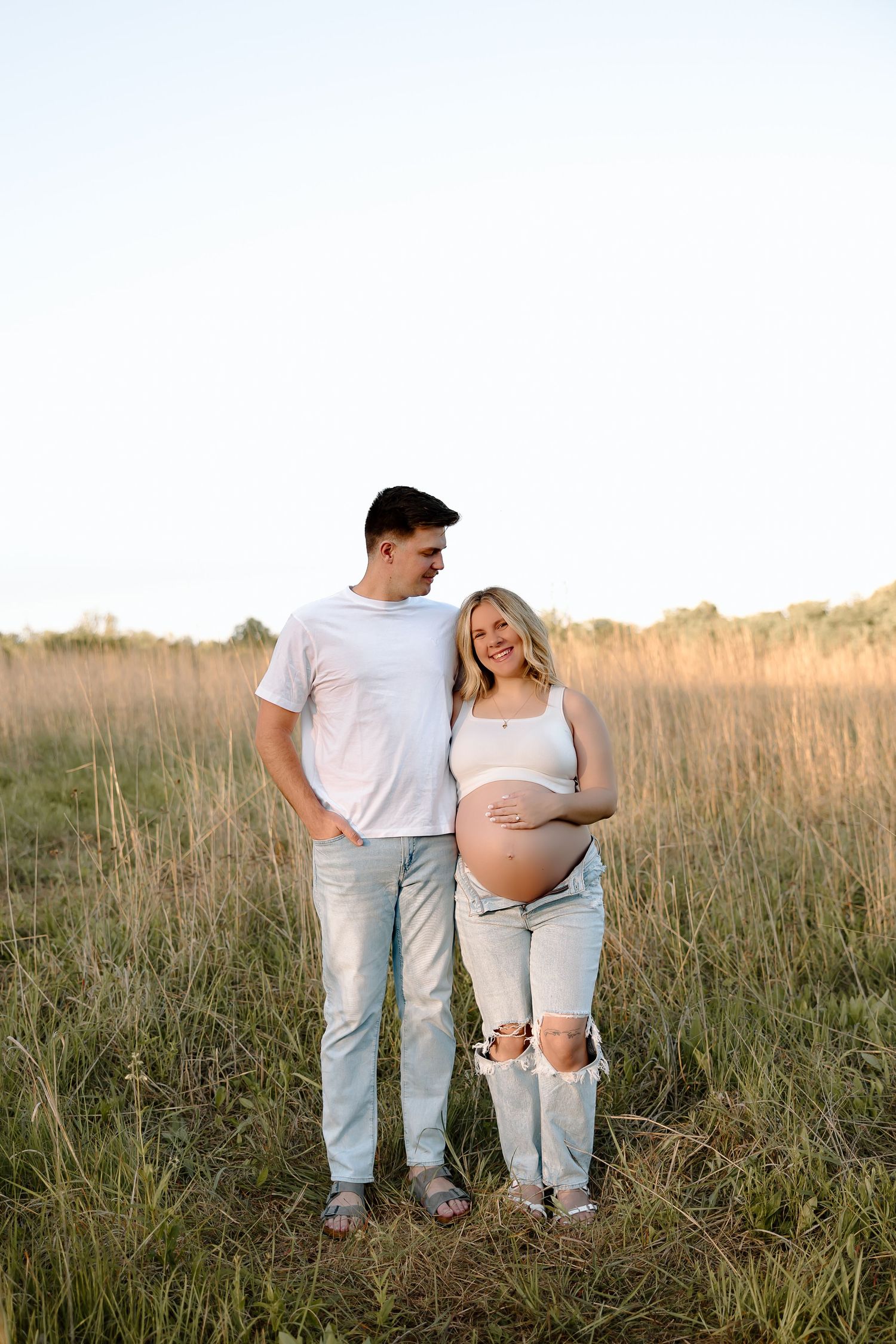 A couple in casual white tops and jeans walk through a golden field during sunset for their maternity photoshoot.