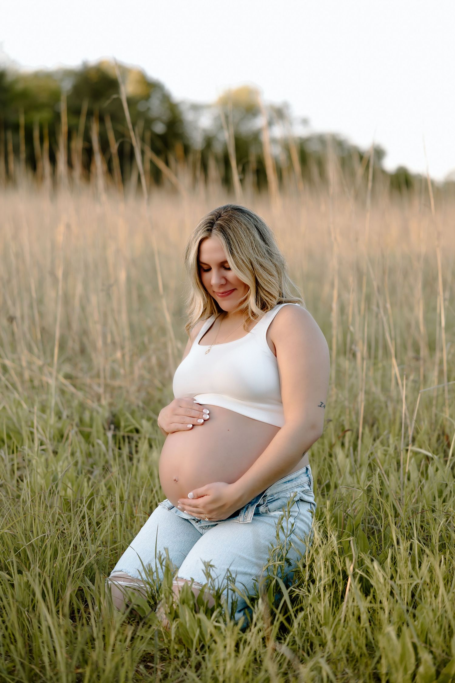 Maternity photo series showing an expectant mother in a white tank top sitting peacefully in a golden grass field.
