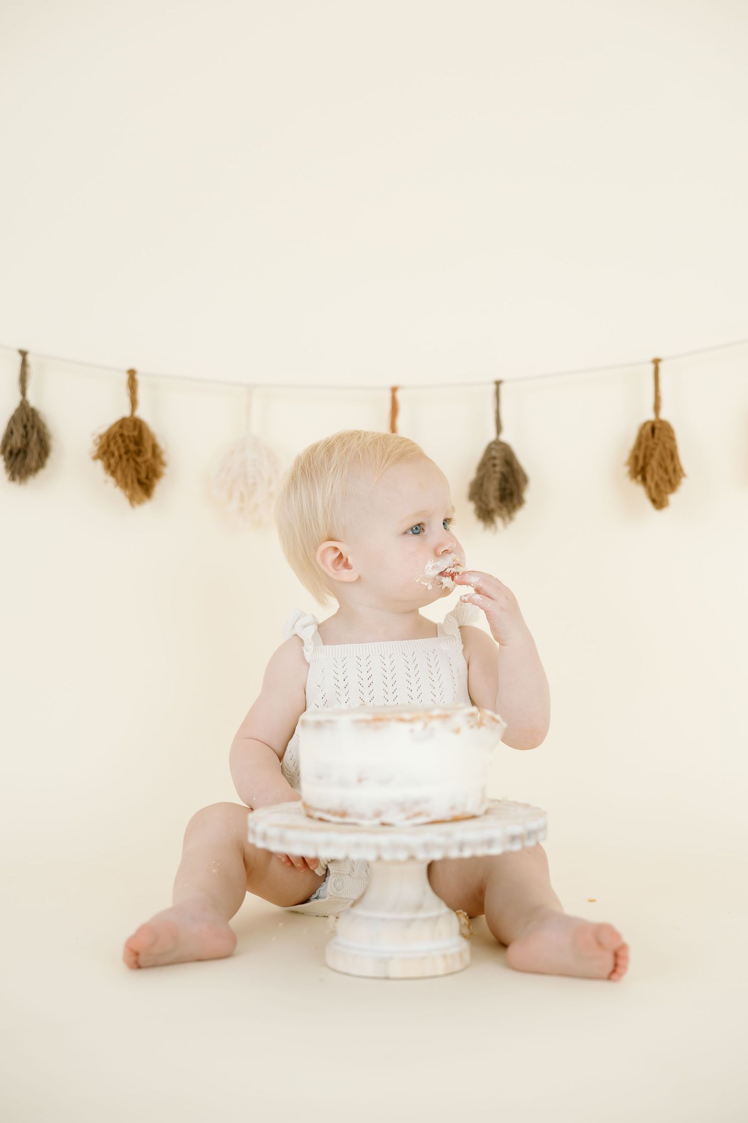 Baby in white diaper reaches for and plays with white birthday cake on cake stand against neutral background.