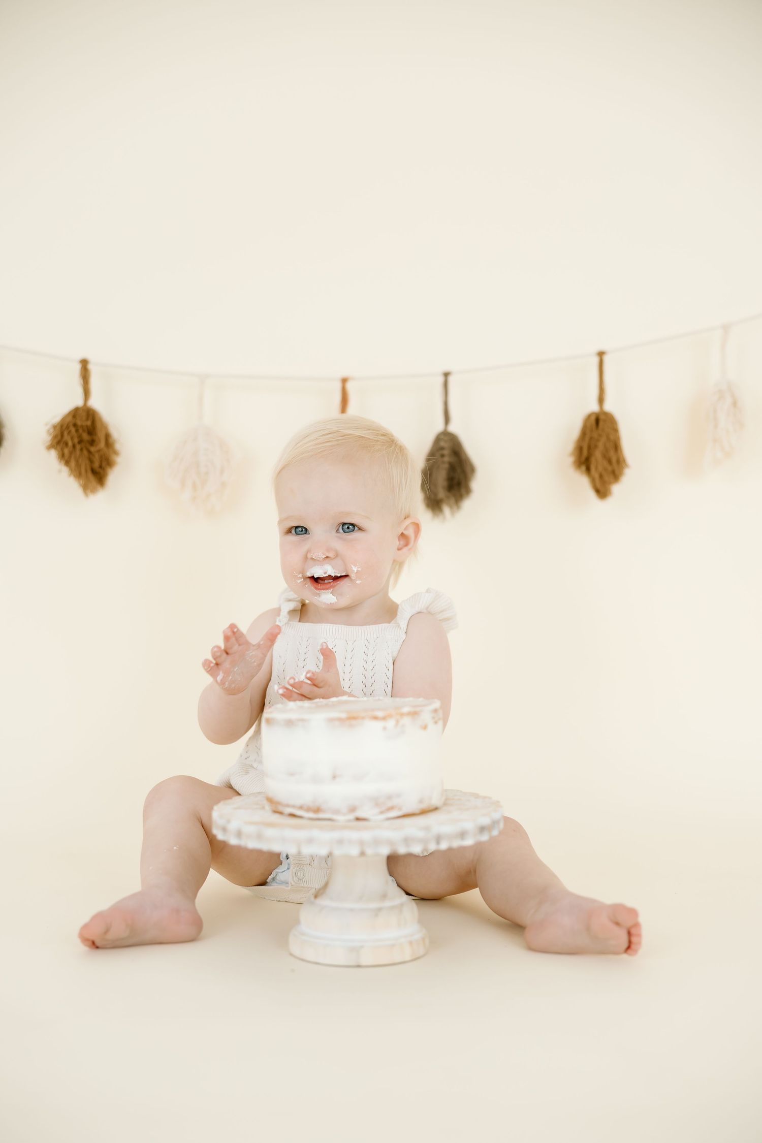 A baby in white sits on a cream backdrop with a white tiered cake during a first birthday cake smash photo session.