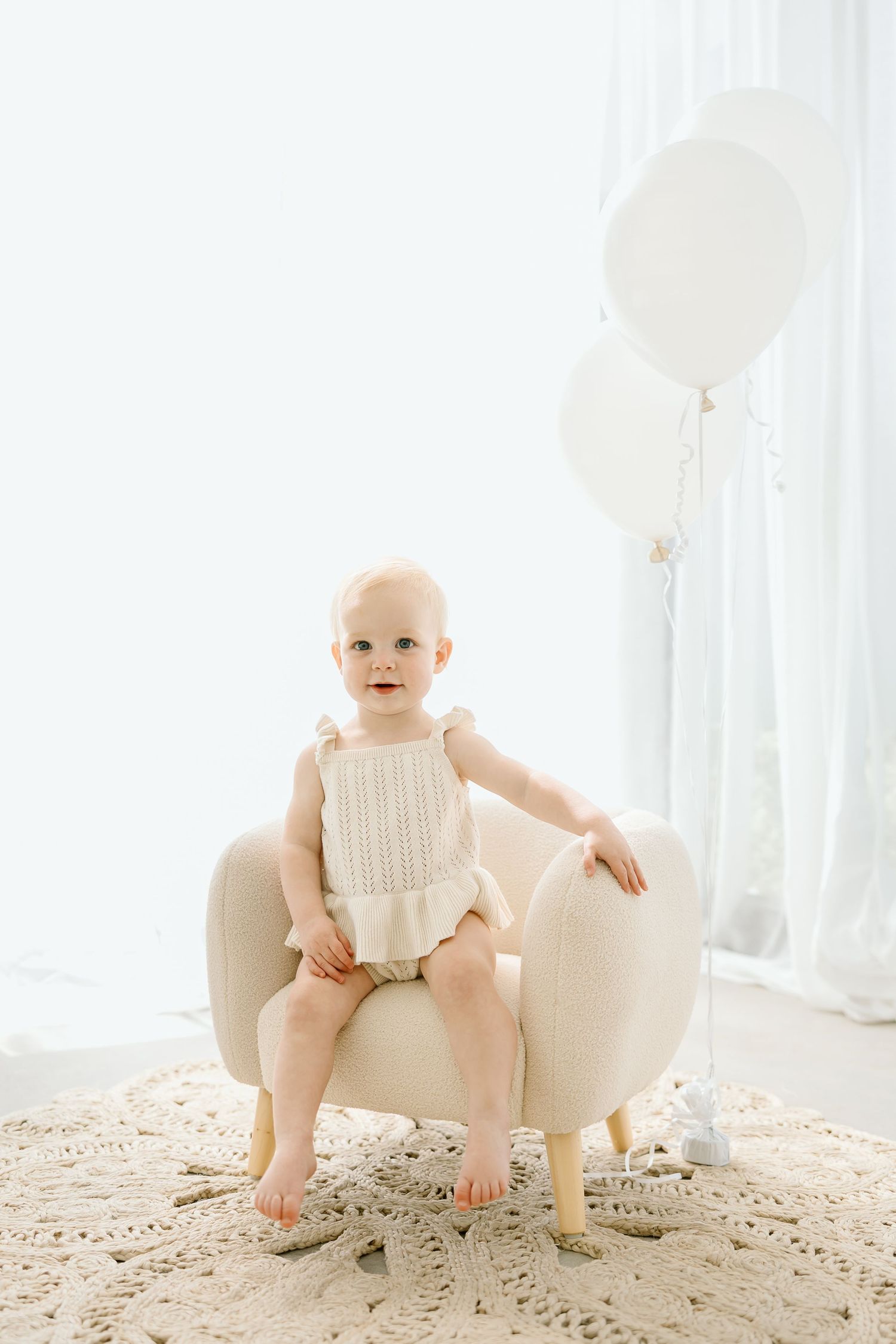 Baby in white knit outfit sitting on cream chair with white balloons in bright studio setting.