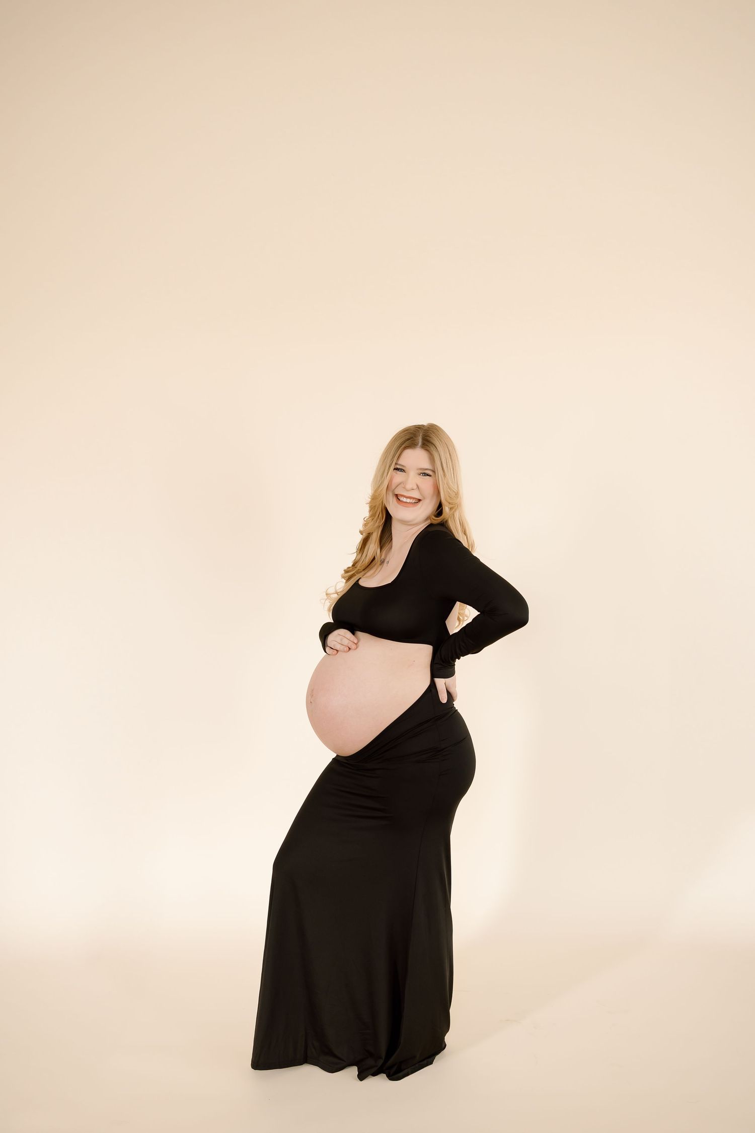 Expectant mother in long black dress poses elegantly against beige background during maternity photoshoot.
