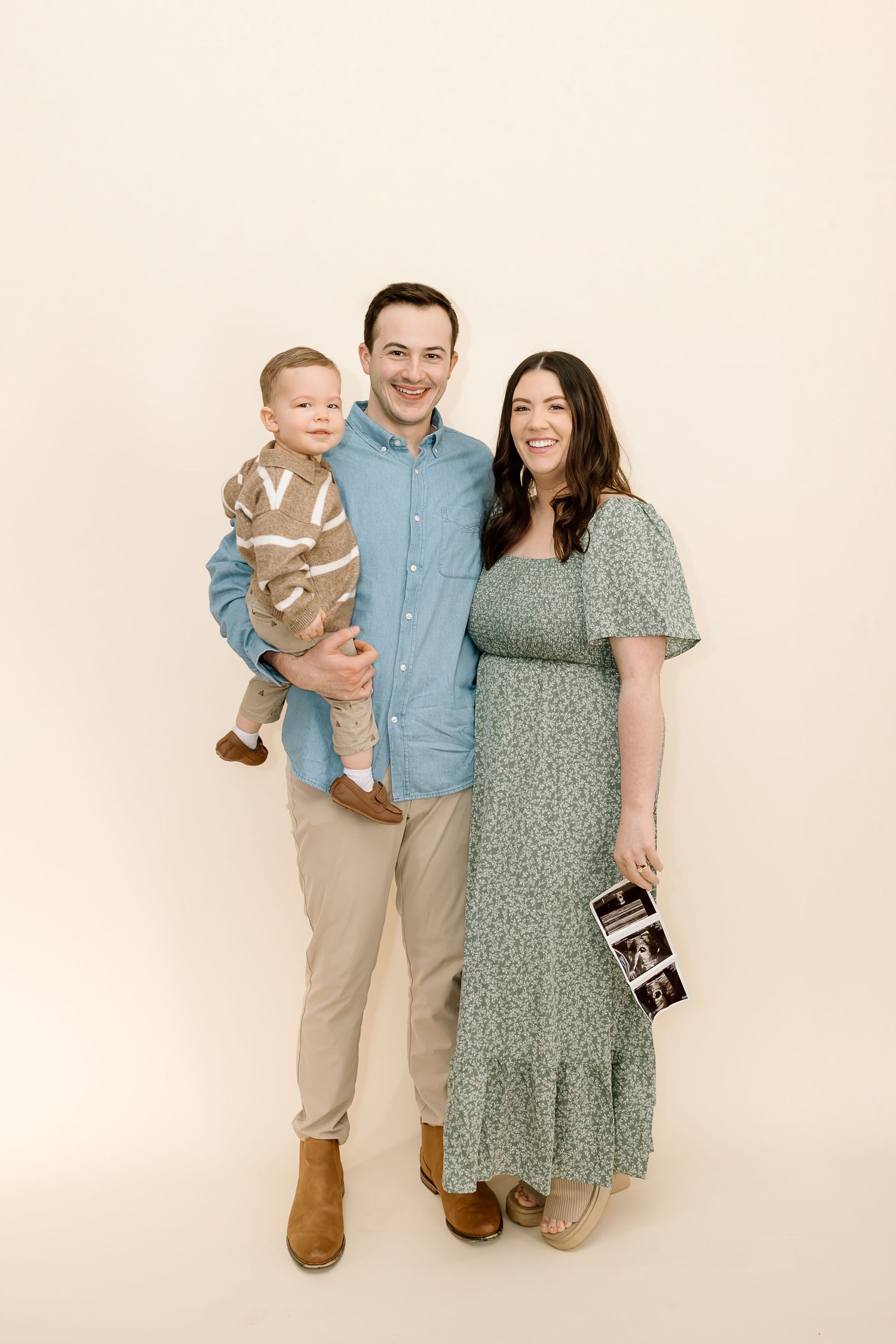 Family of three poses together against cream backdrop while holding ultrasound photo in maternity announcement.