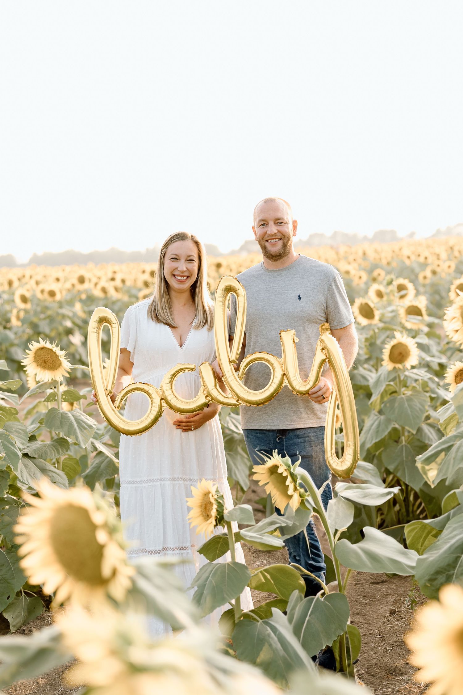 A couple stands together in a golden sunflower field at sunset holding a balloon that spells 'boy'.