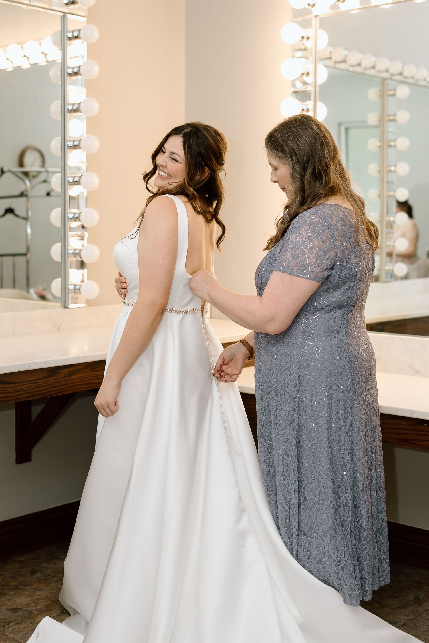 A bride in a white wedding dress gets help with final adjustments from someone wearing a grey lace gown in a dressing room.