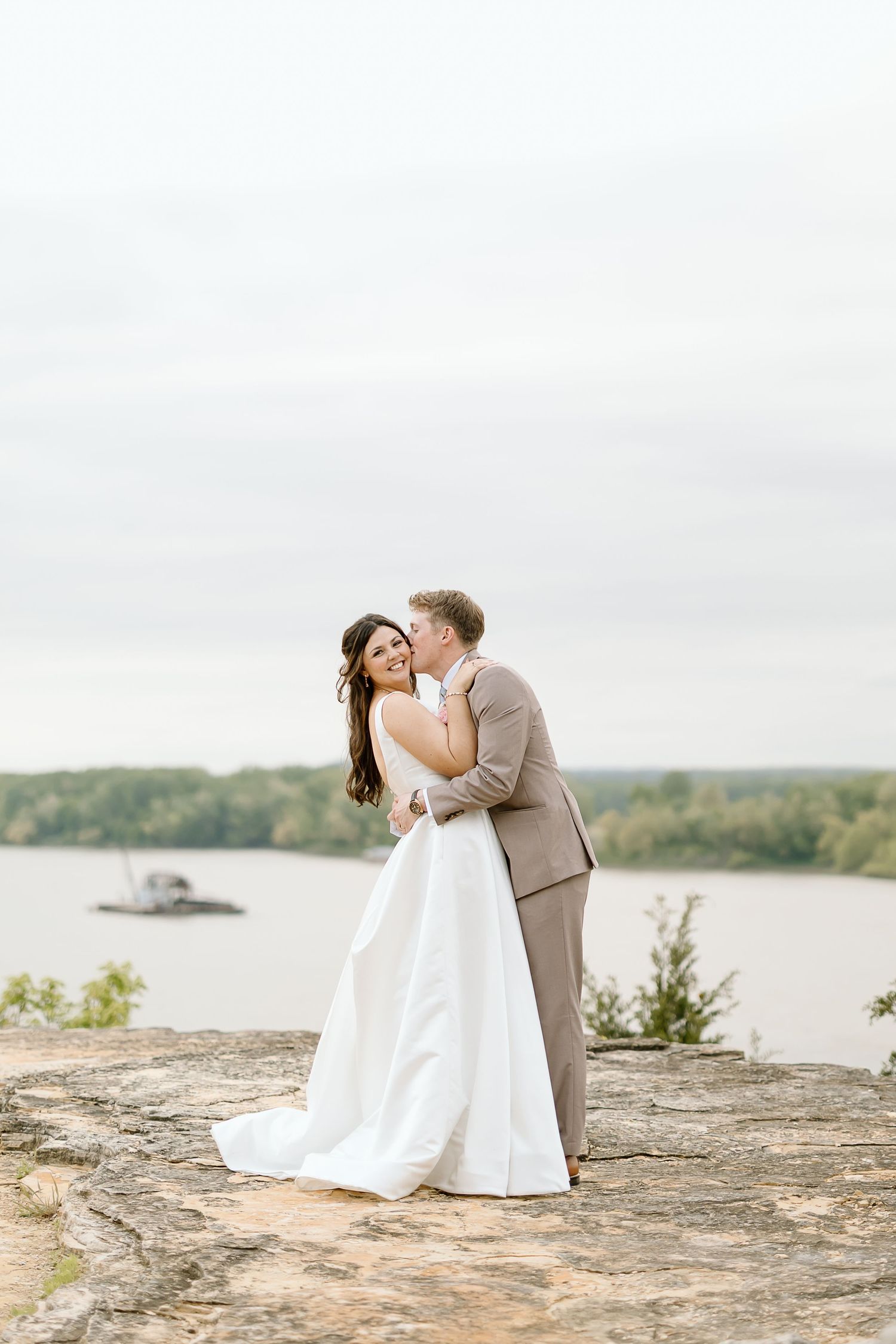 A couple shares a romantic moment on a rocky cliff overlooking a serene lake on their wedding day.