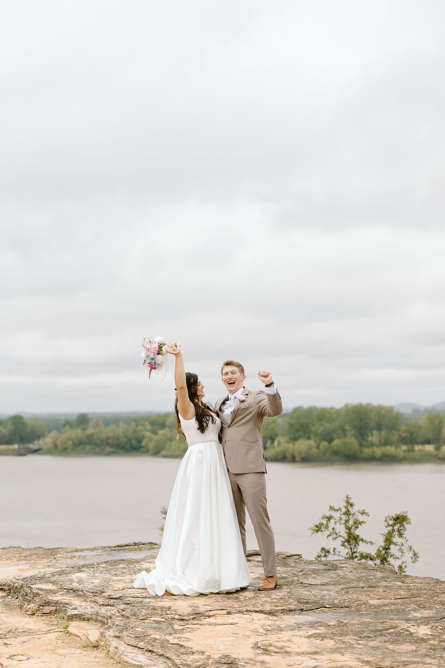 Newlywed couple celebrates joyfully on a cliff overlooking a scenic lake and forest landscape on their wedding day.