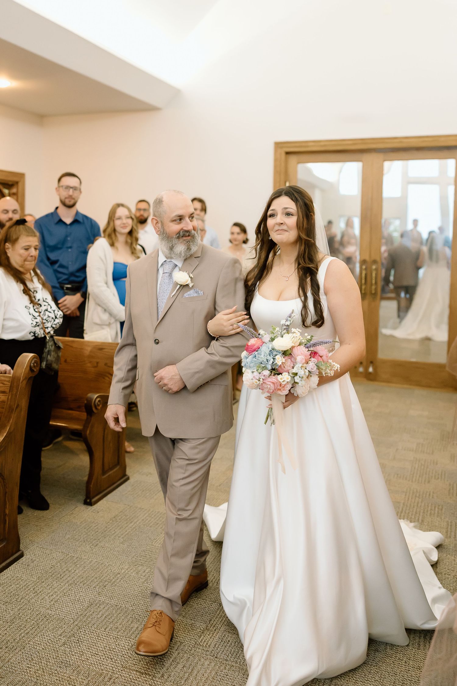 A bride in a white wedding gown walks down the church aisle holding a pink and white bouquet.