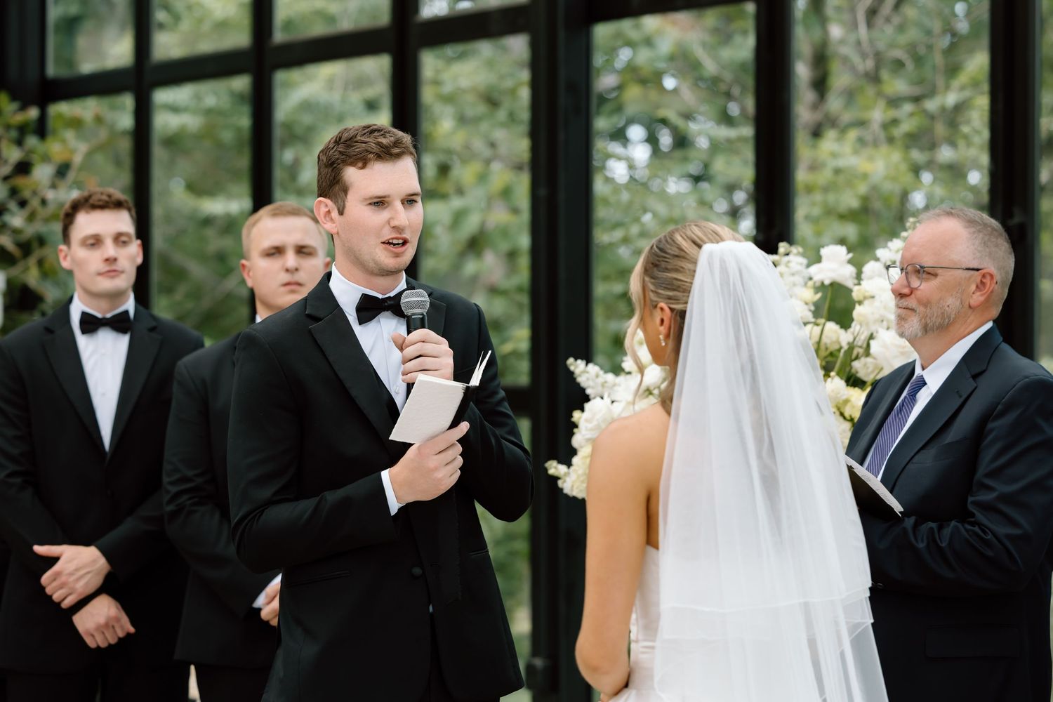 Groom reads wedding vows at altar with groomsmen and officiant during elegant indoor ceremony.