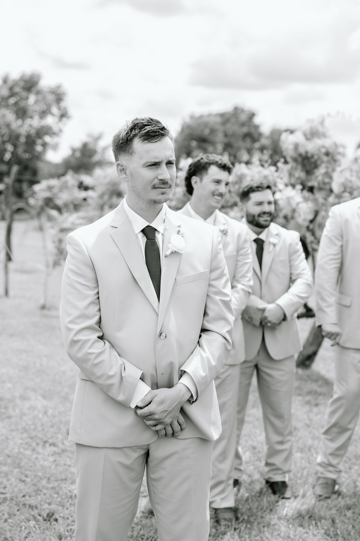 Groom in light suit stands at altar with groomsmen lined up behind him outdoors at a wedding ceremony.