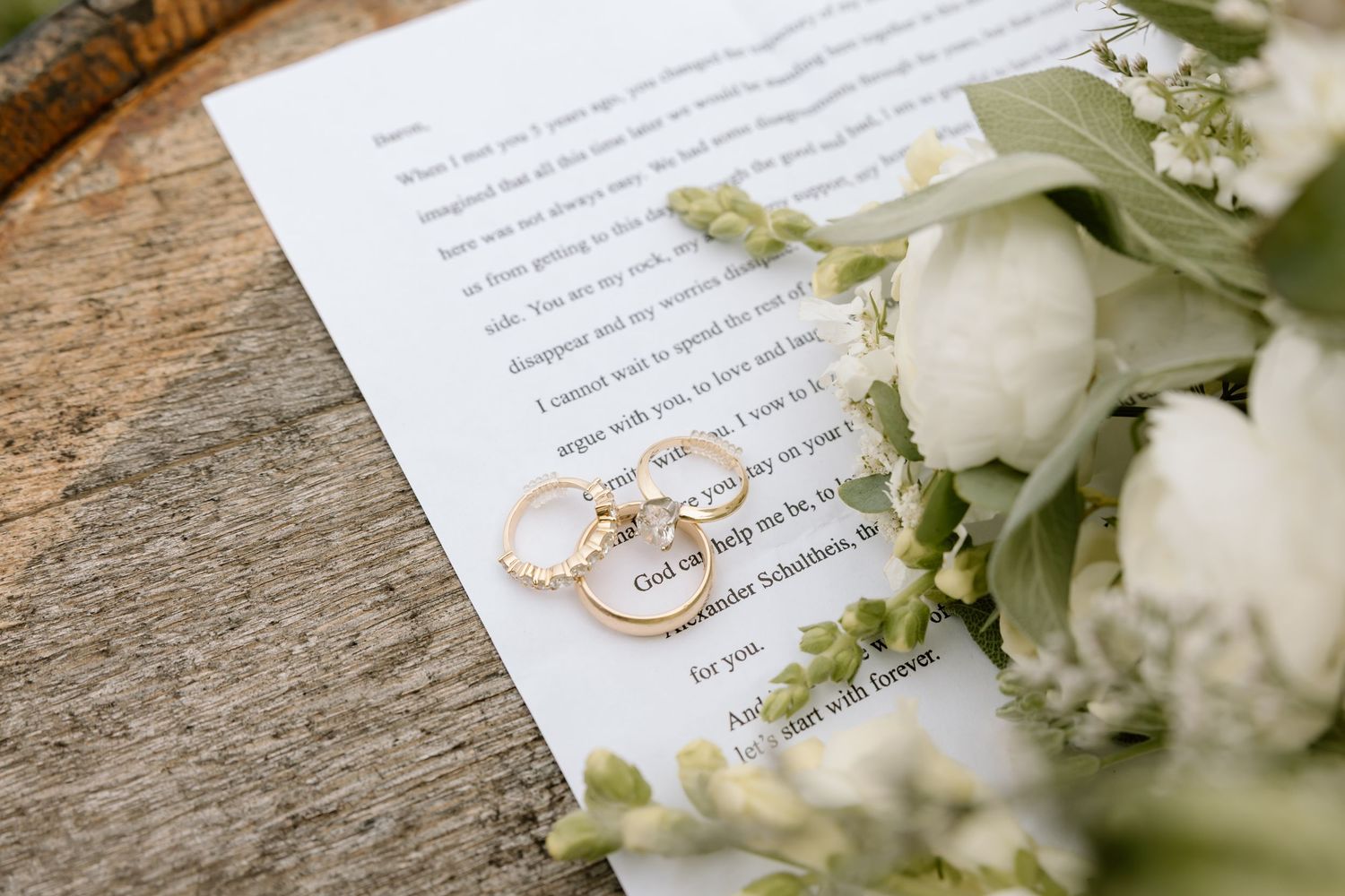 Gold wedding rings rest on handwritten vows beside a white floral bouquet with roses and greenery on a wooden surface.