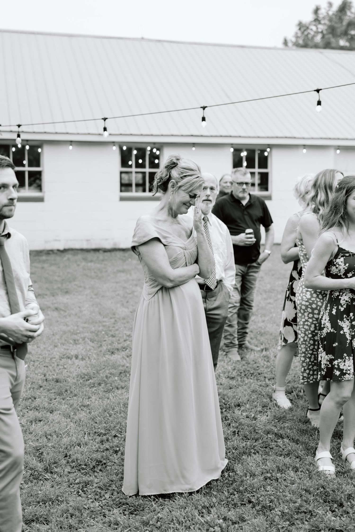 Black and white photo of wedding guests in formal attire gathered on a lawn outside a white barn venue with string lights.