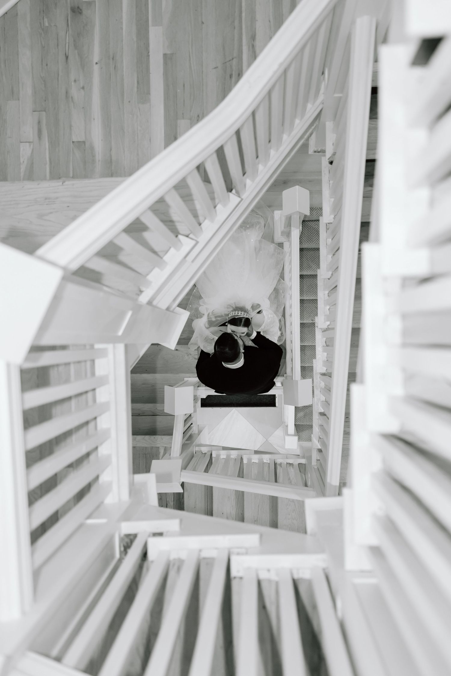 Black and white aerial view of a couple embracing on a winding staircase with white railings and wooden floors.