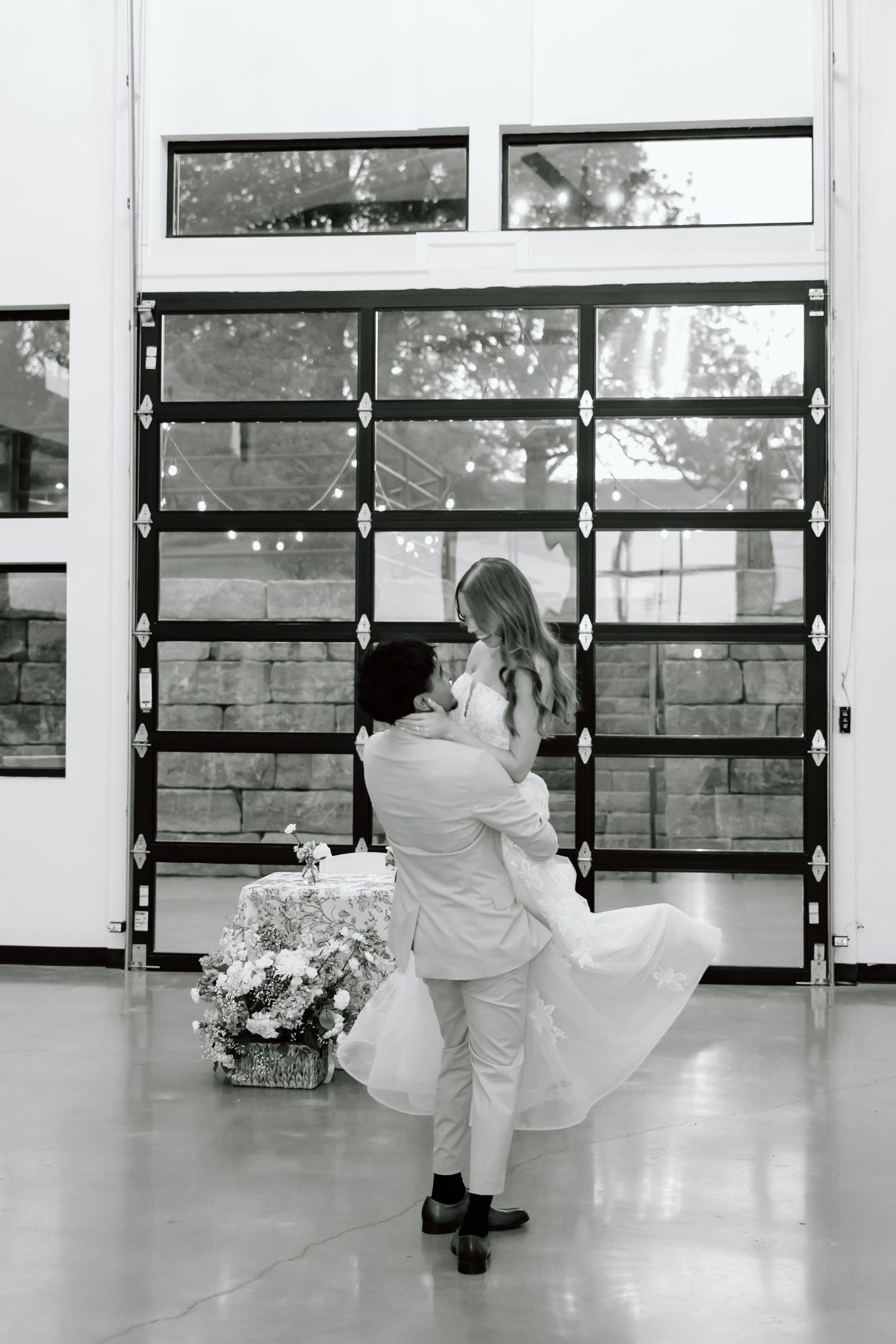 Black and white photo of a groom lifting his bride in a white dress near a large glass garage door at their wedding.