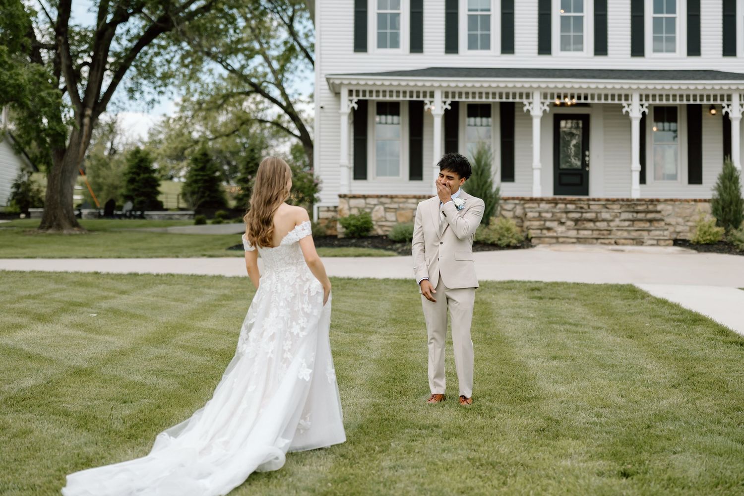 A bride in a white gown and groom in a tan suit share a first look moment on a green lawn before a white farmhouse.
