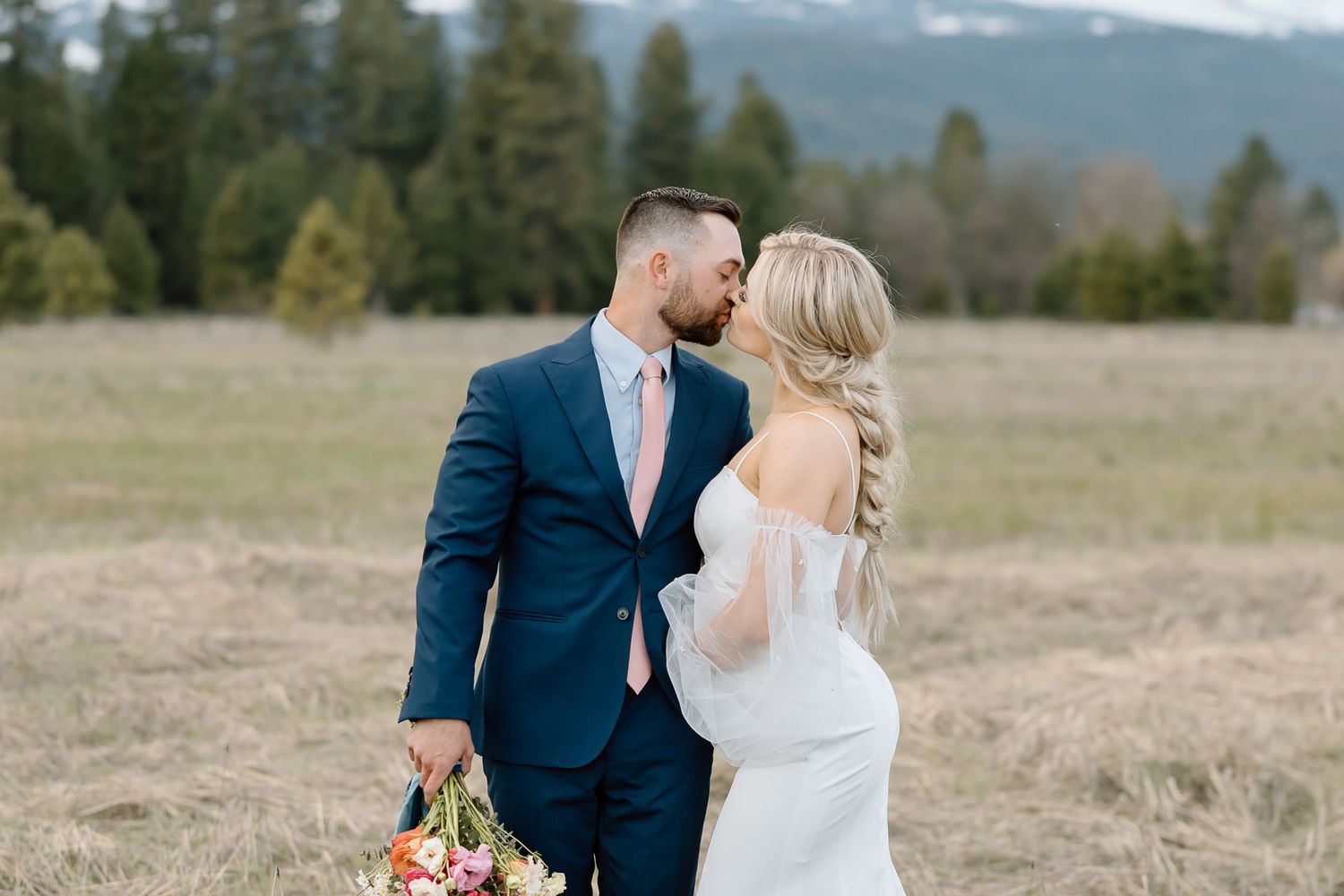 Intimate moment between wedding couple in field with mountains and forest backdrop.