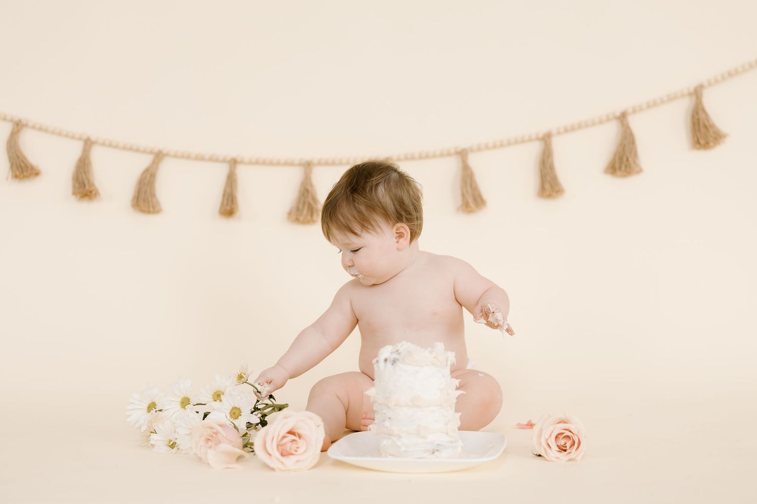 Baby sitting with white cake and floral decorations against neutral backdrop with tassels.