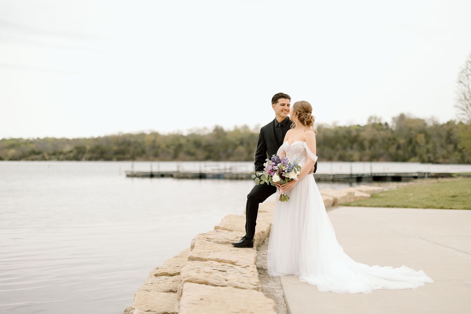 Newlyweds pose on stone wall by lake during sunset wedding photos.