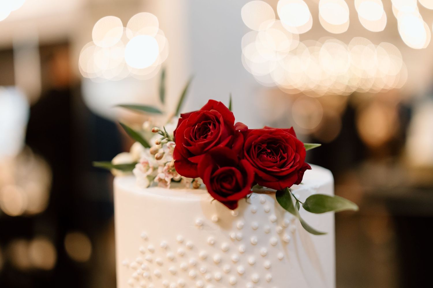 Three tiered white wedding cake decorated with red roses and gold accents sits elegantly on display.