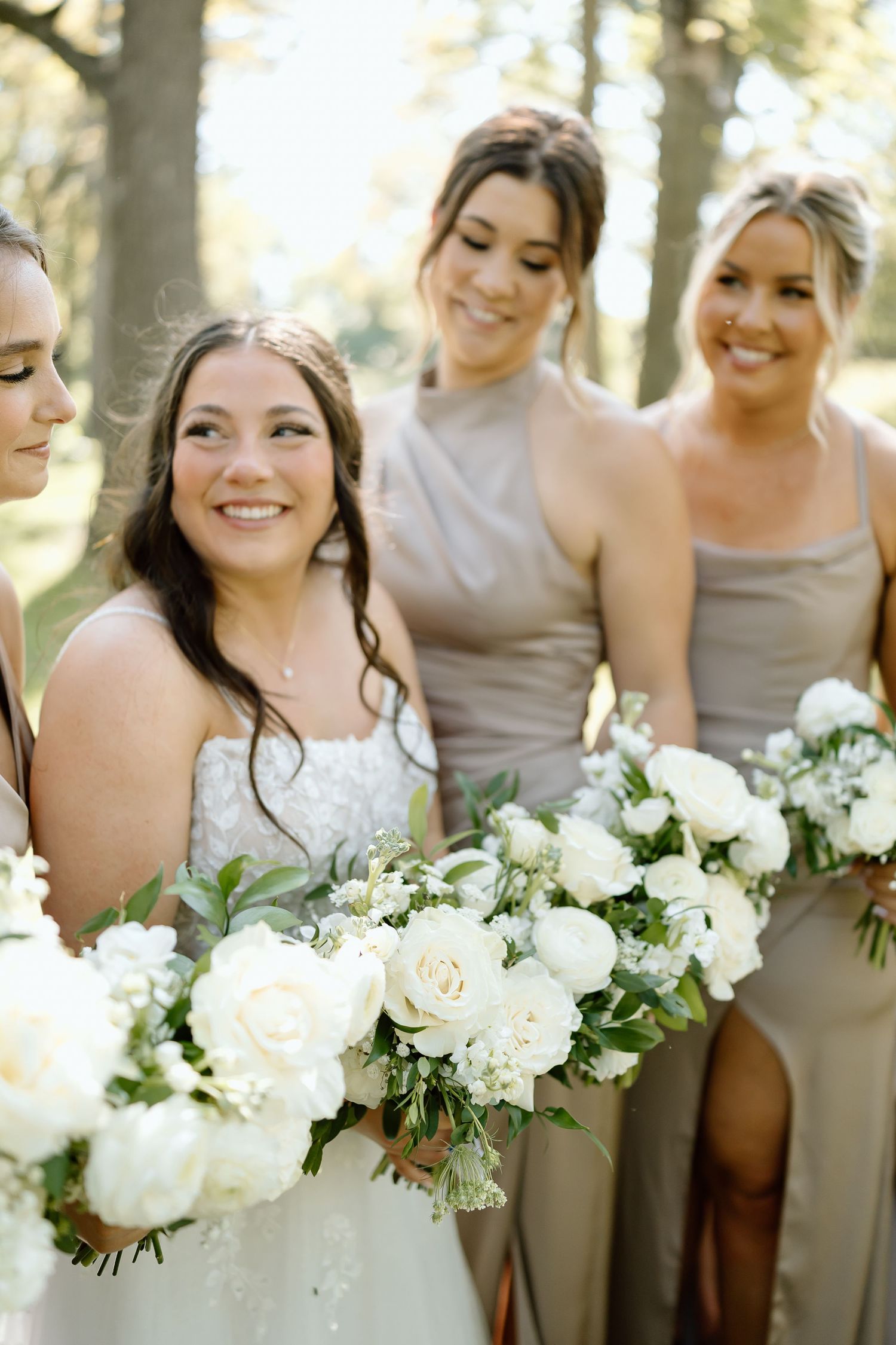 Bridesmaids in taupe dresses hold elegant white and green bouquets while smiling in natural outdoor lighting.
