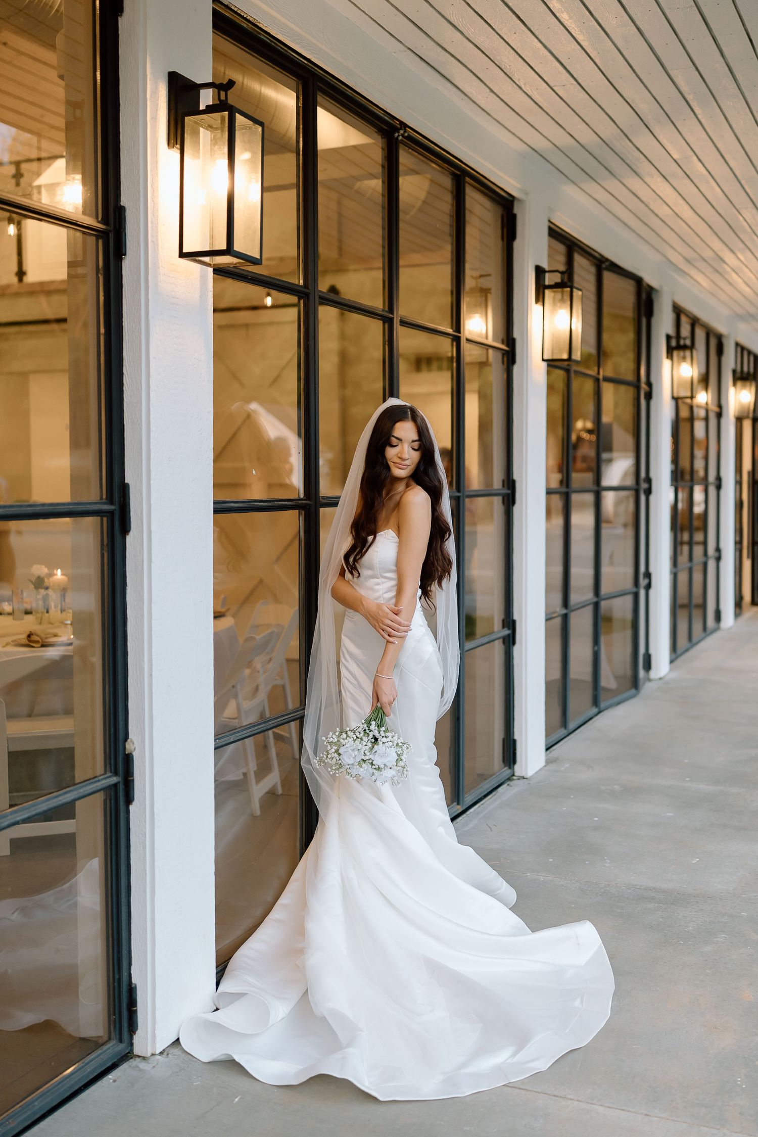 A bride in a flowing white wedding dress poses elegantly in a modern hallway with glass windows and lantern lighting.
