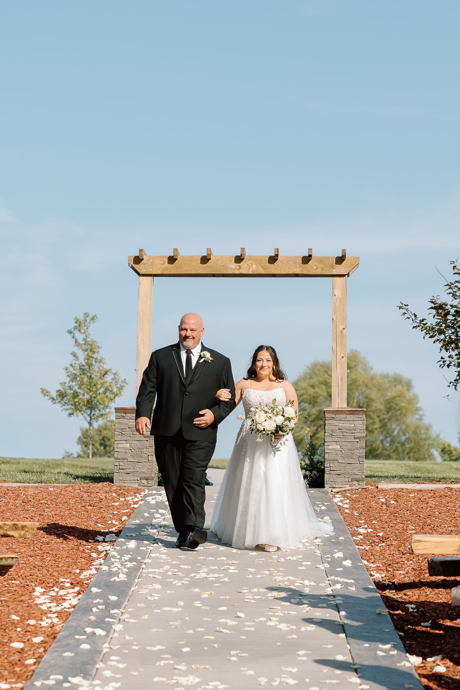 A couple walks down a petal-strewn path under a wooden arch on their wedding day against a blue sky.