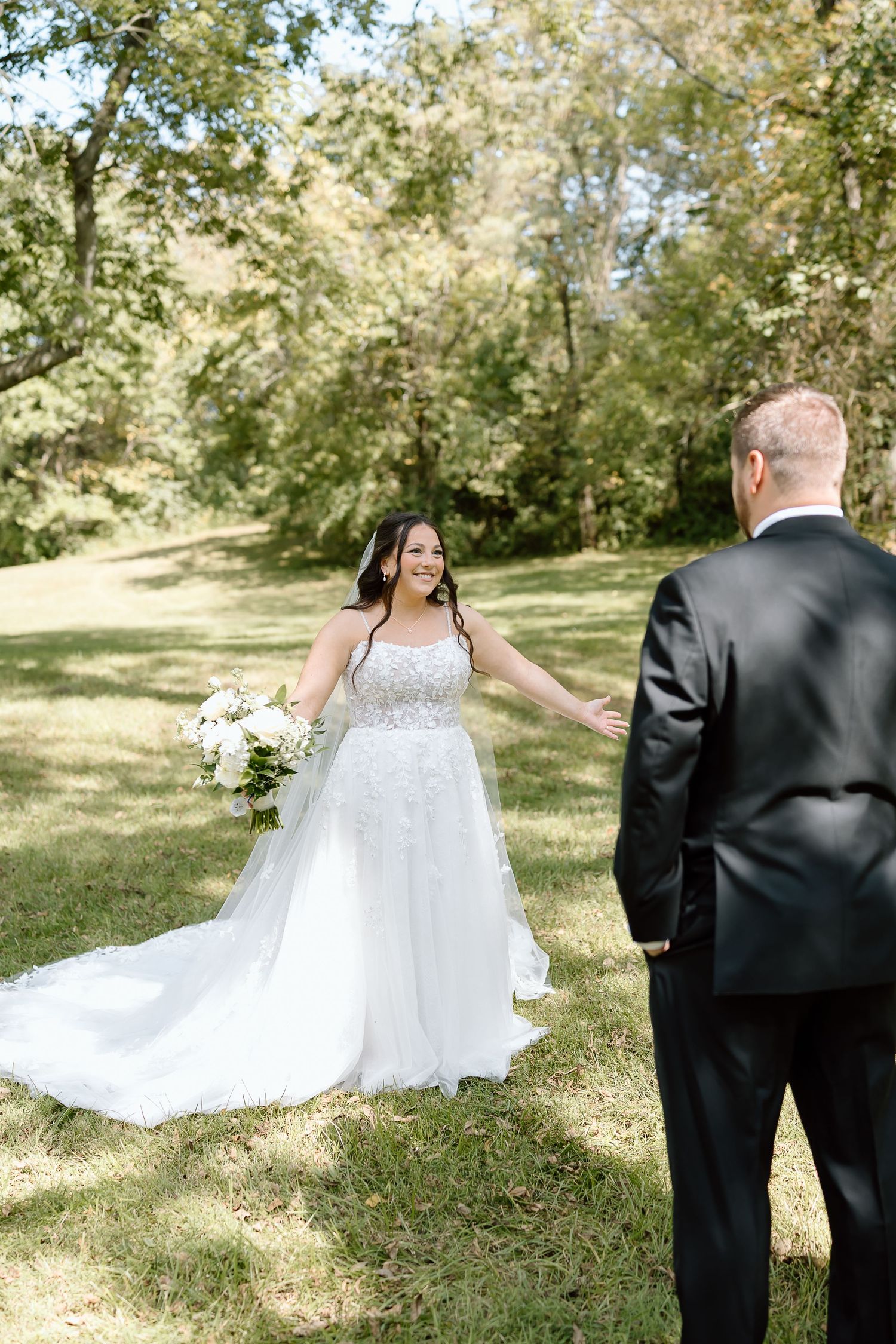 A bride in a flowing white dress twirls joyfully in a sun-dappled garden setting while someone in a black suit looks on.