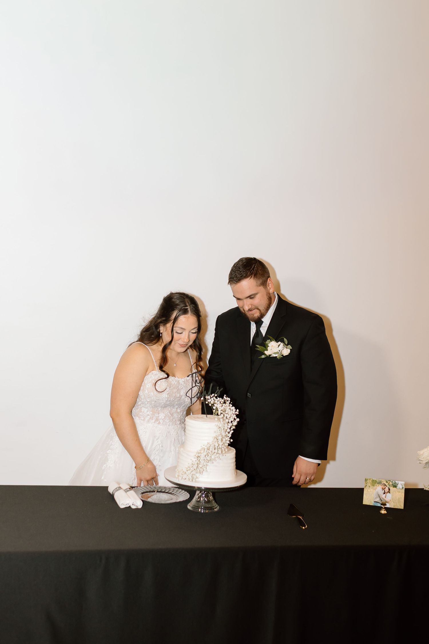 A couple in wedding attire leans over a table with a white tiered wedding cake during their reception celebration.