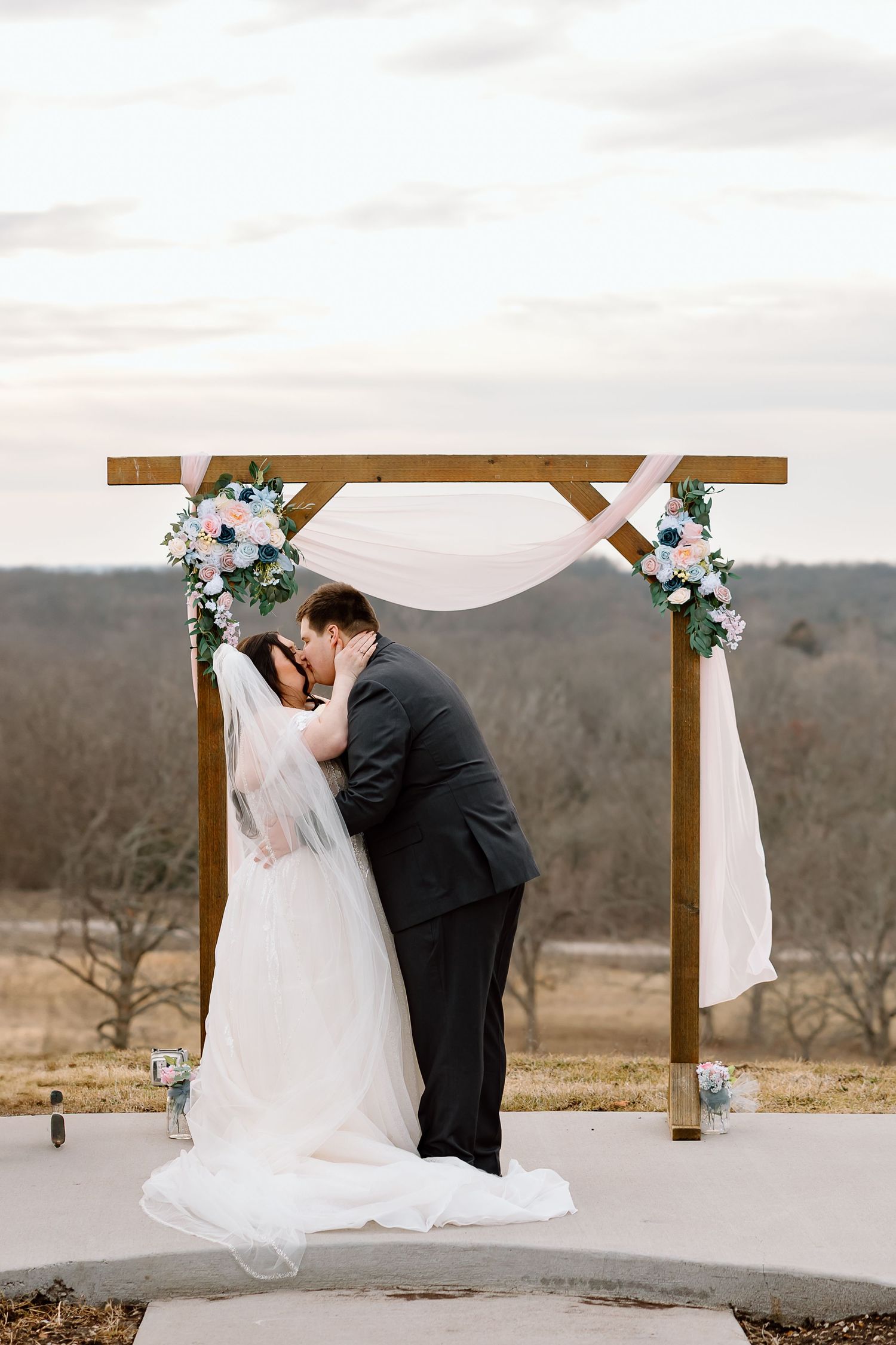 A romantic wedding ceremony moment at a rustic wooden arch decorated with white fabric and flowers against a rural landscape.