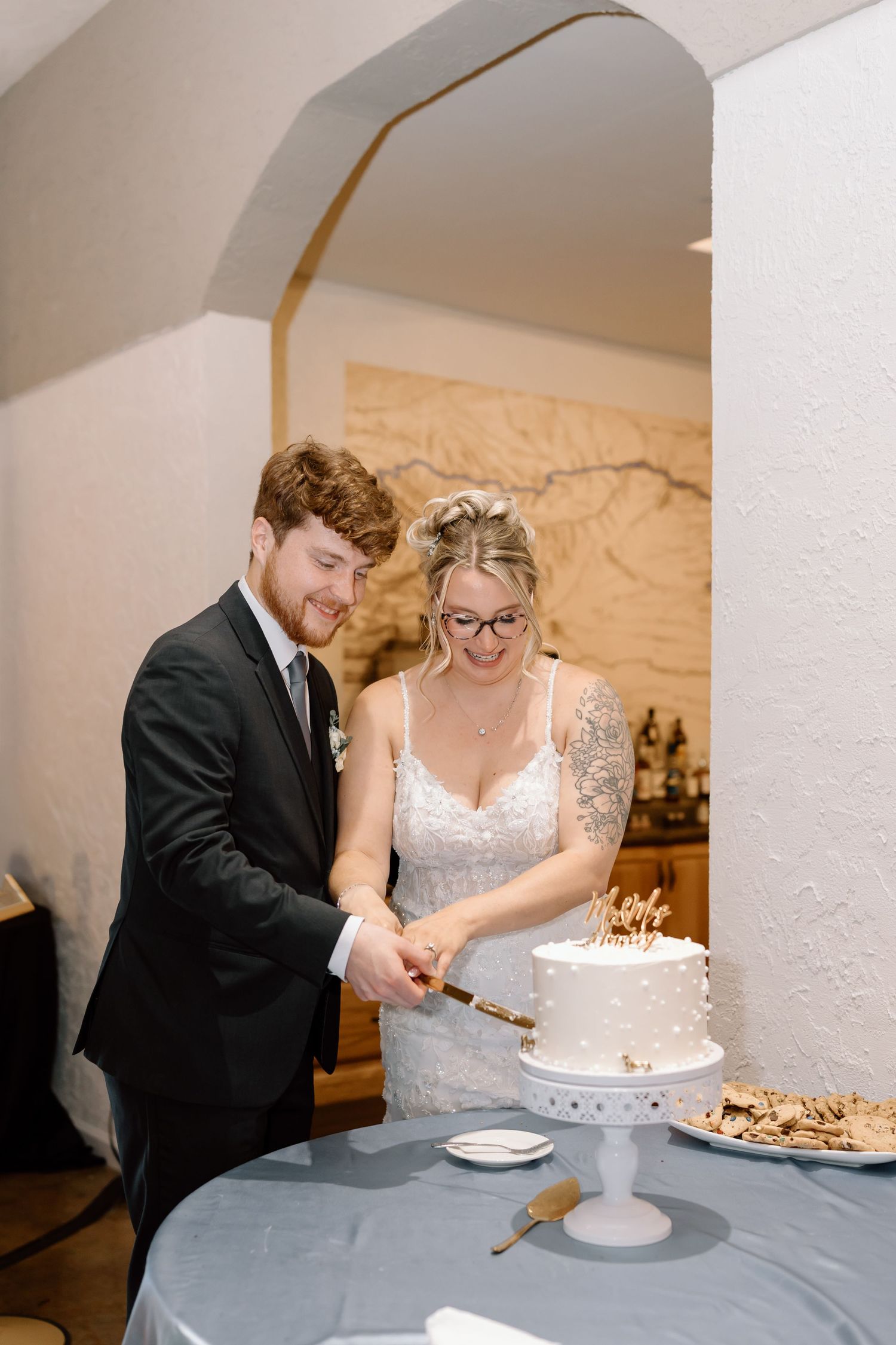 A couple in wedding attire cuts into their white wedding cake together at their reception celebration.