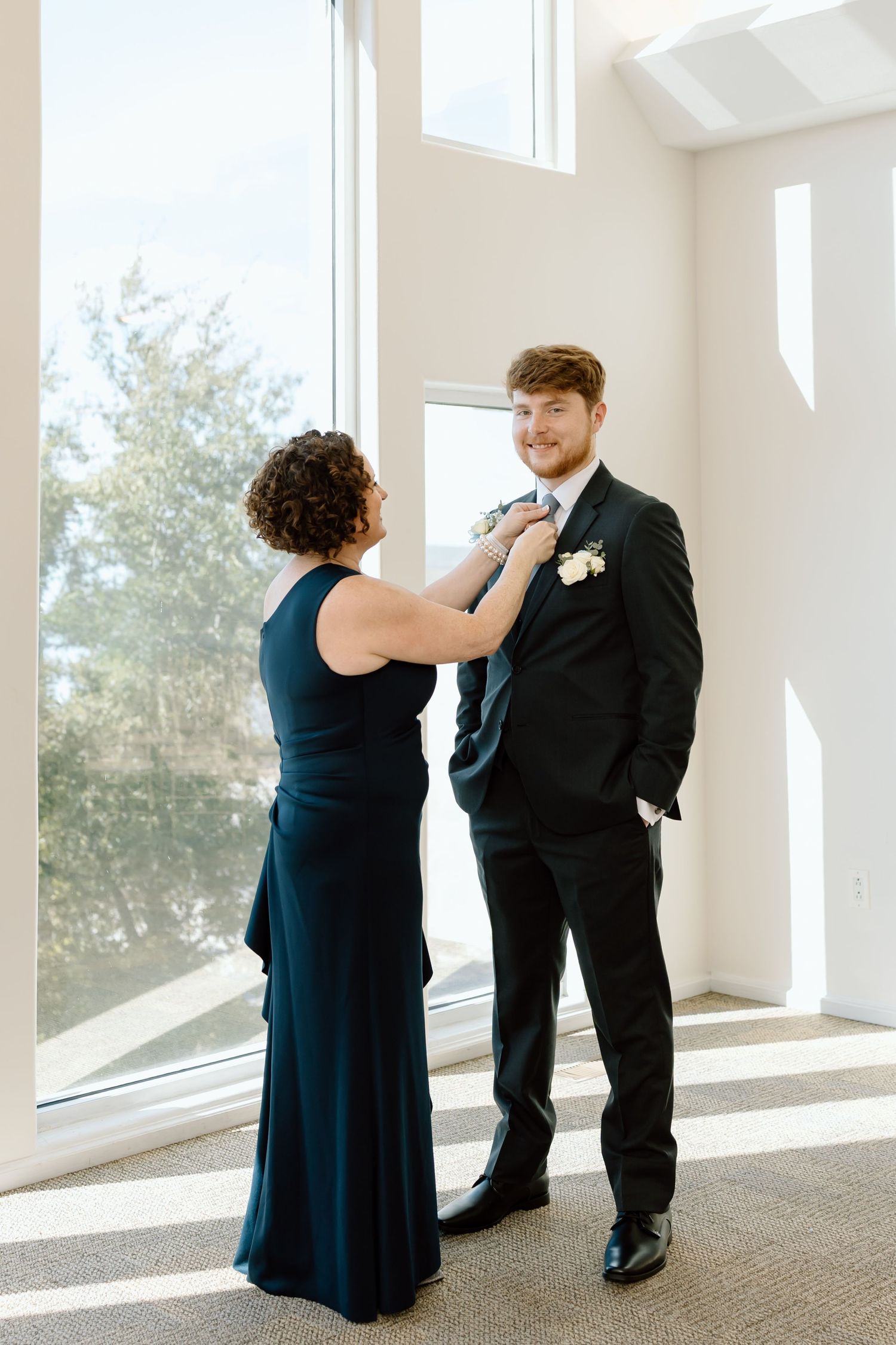 A formal moment in a bright room with a person in navy dress adjusting a tie on someone in a black suit.