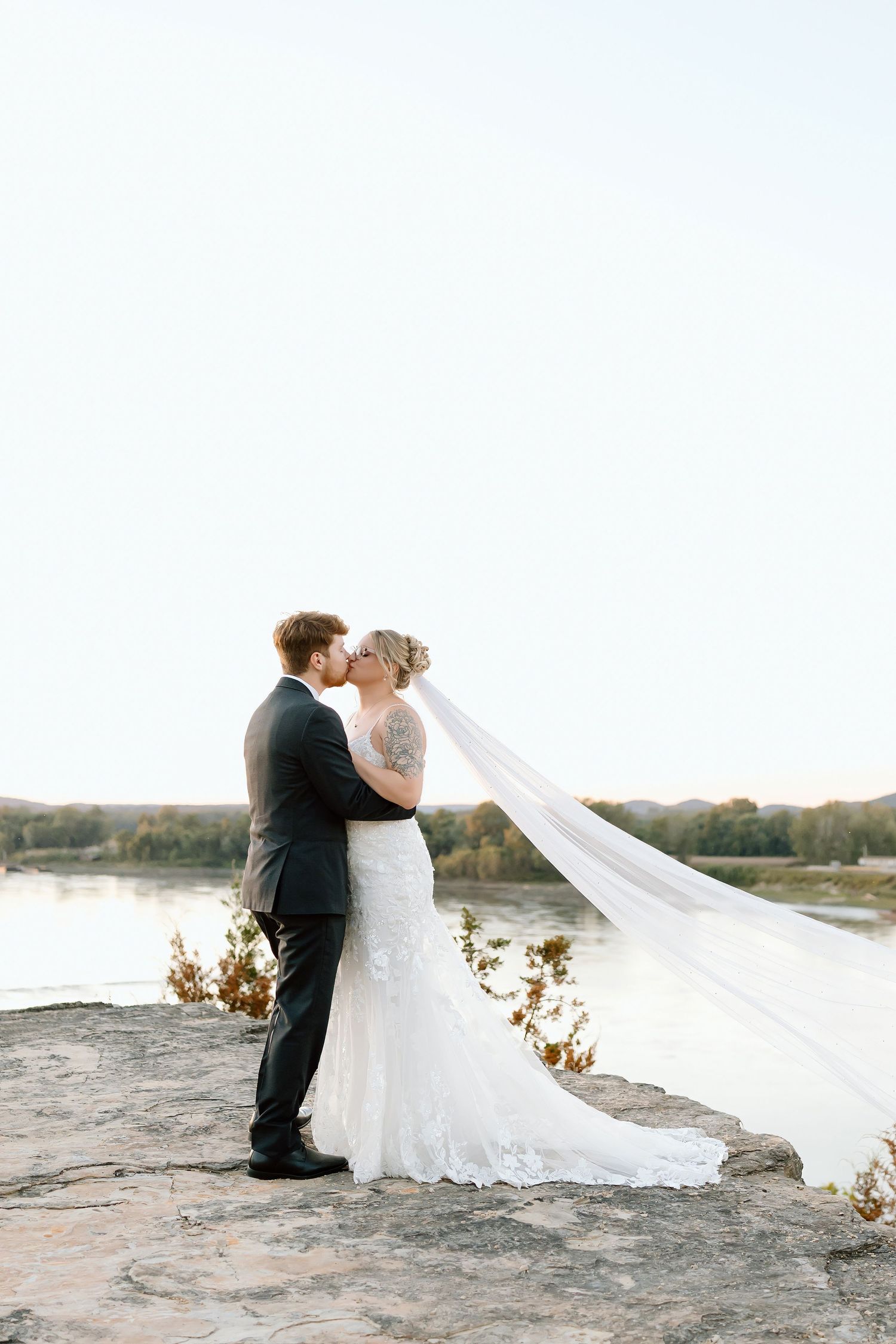 A couple in wedding attire sits on a rocky outcrop as a long veil flows dramatically in the wind at sunset.