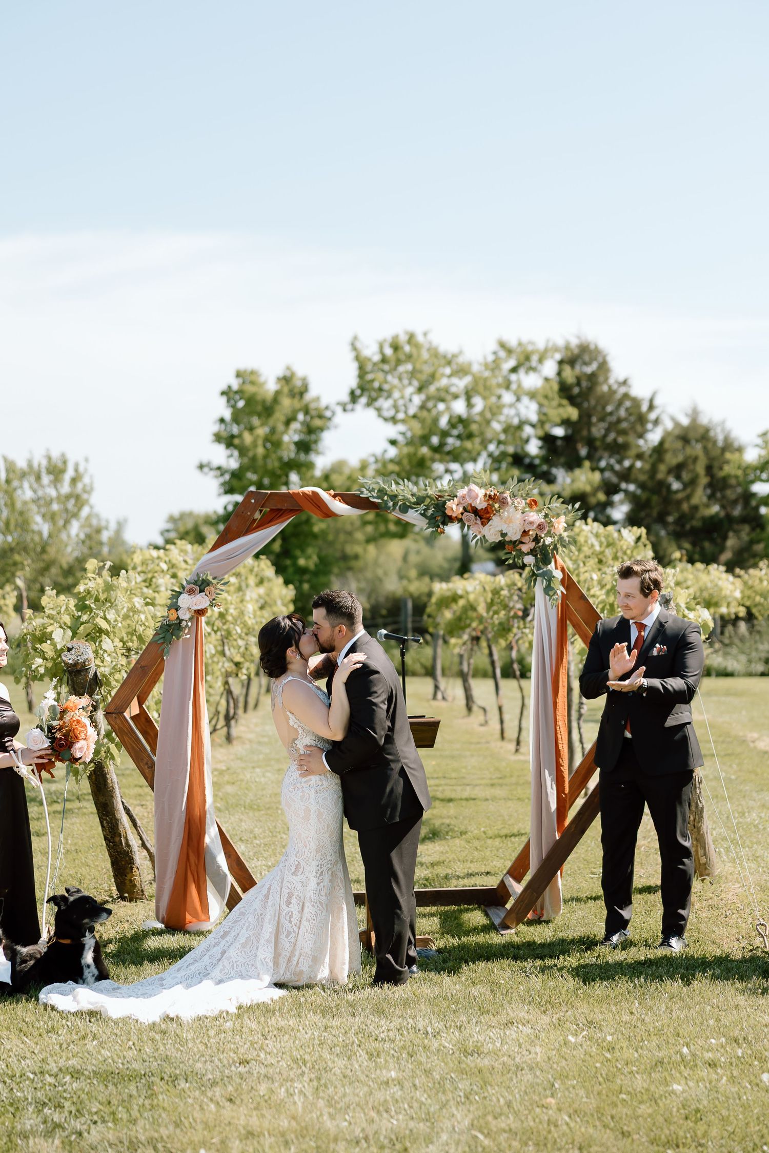 Outdoor wedding ceremony under a wooden arch decorated with white flowers and fabric on a sunny day.