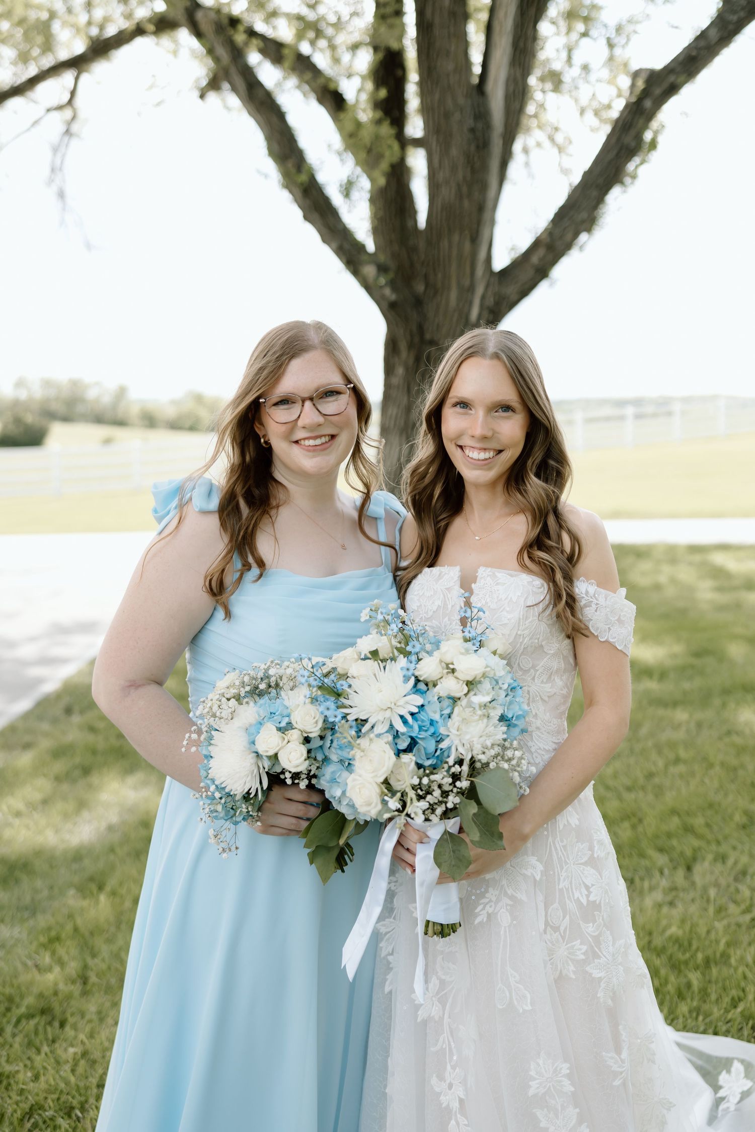 Bride in lace off-shoulder gown holds blue and white bouquet beside bridesmaid in light blue dress.