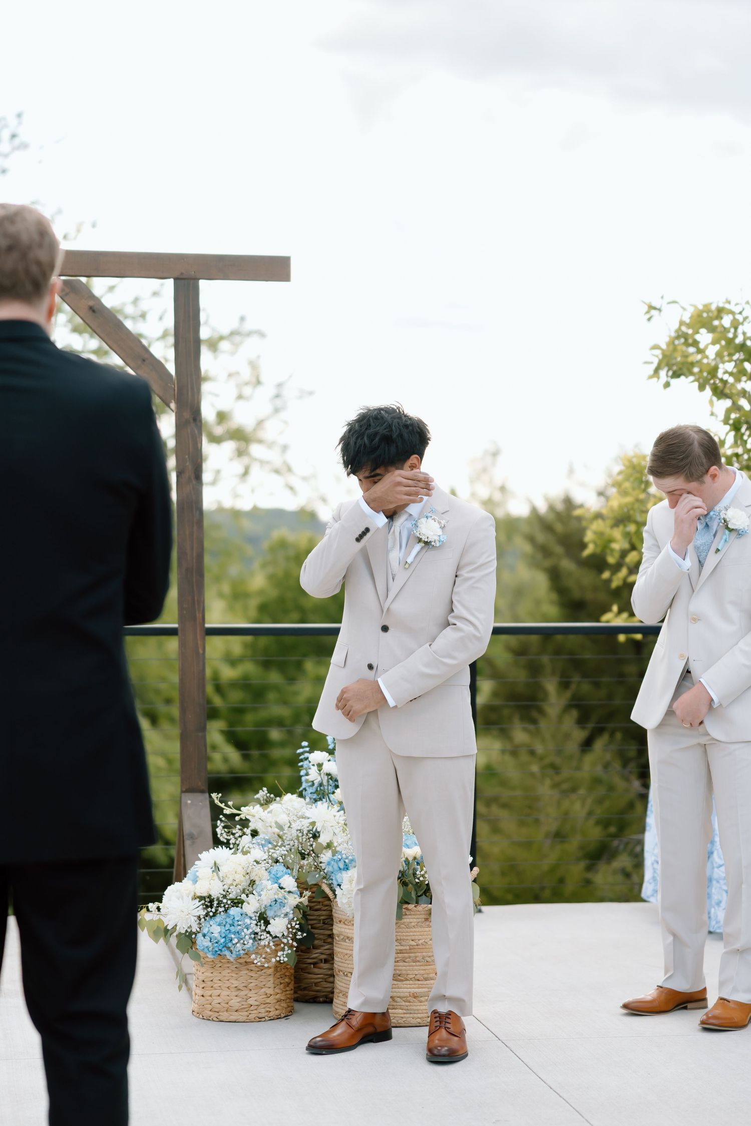 Emotional groom crying at outdoor wedding altar wearing beige suit with boutonniere, surrounded by groomsmen and floral arrangements.