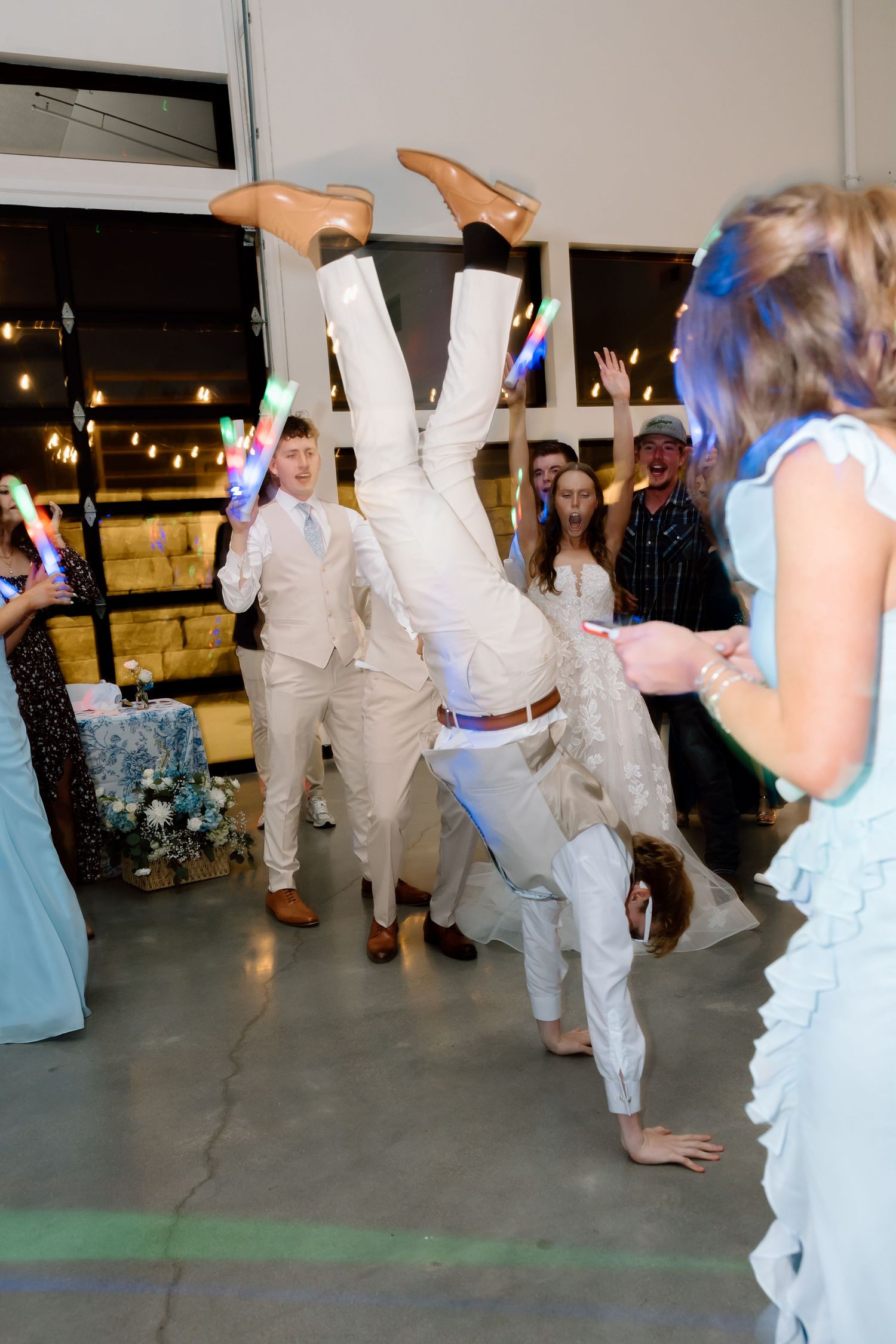 A wedding guest performs a headstand on the dance floor while others celebrate with glow sticks at a festive reception.