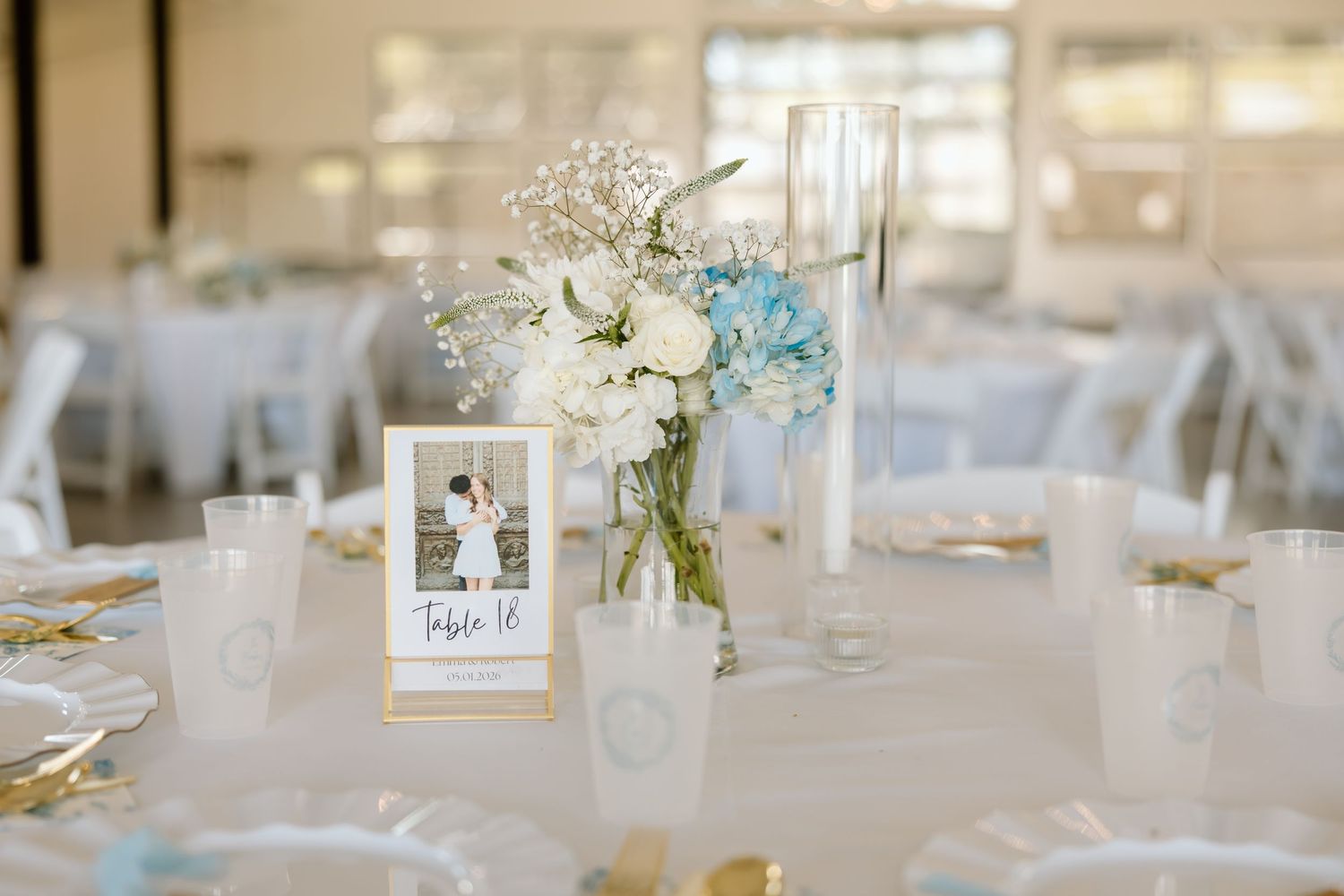 Elegant wedding reception table with white and blue floral centerpiece, gold accents, and a framed couples photo table number.