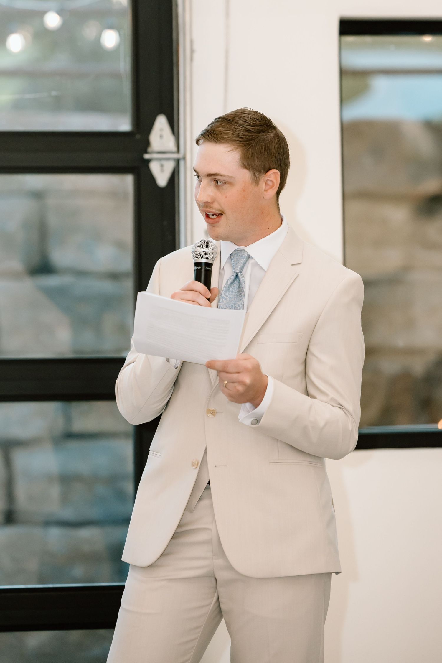 A groomsman in a beige suit delivers a heartfelt wedding speech holding a microphone and notes.