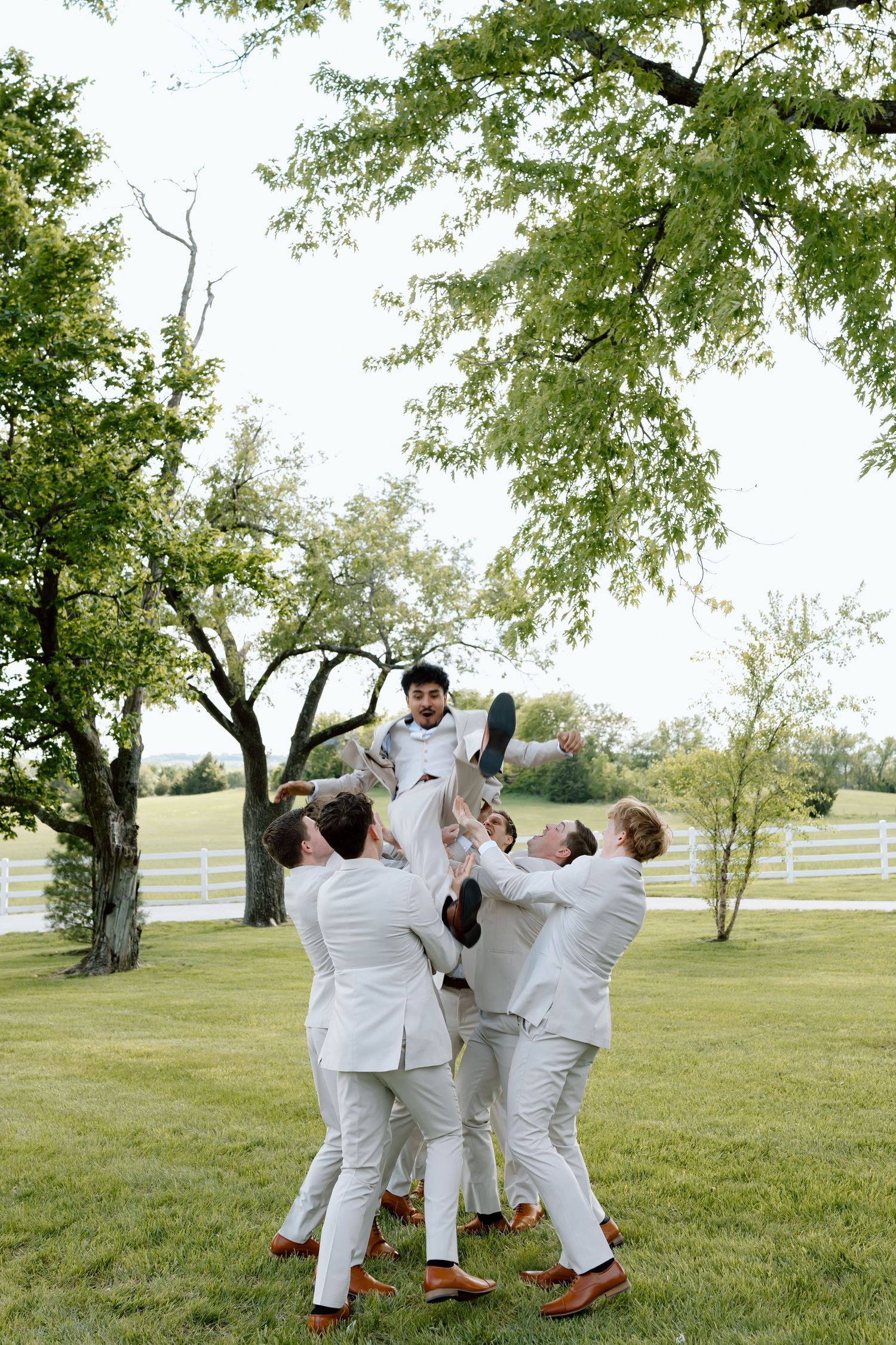 Groomsmen in white suits tossing the groom into the air outdoors on a green lawn at a wedding celebration.