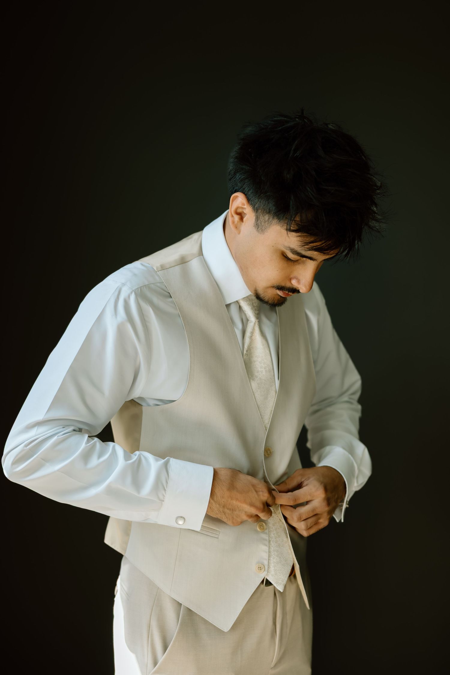 Groom in white suit and tie buttoning his vest while looking down against dark background.