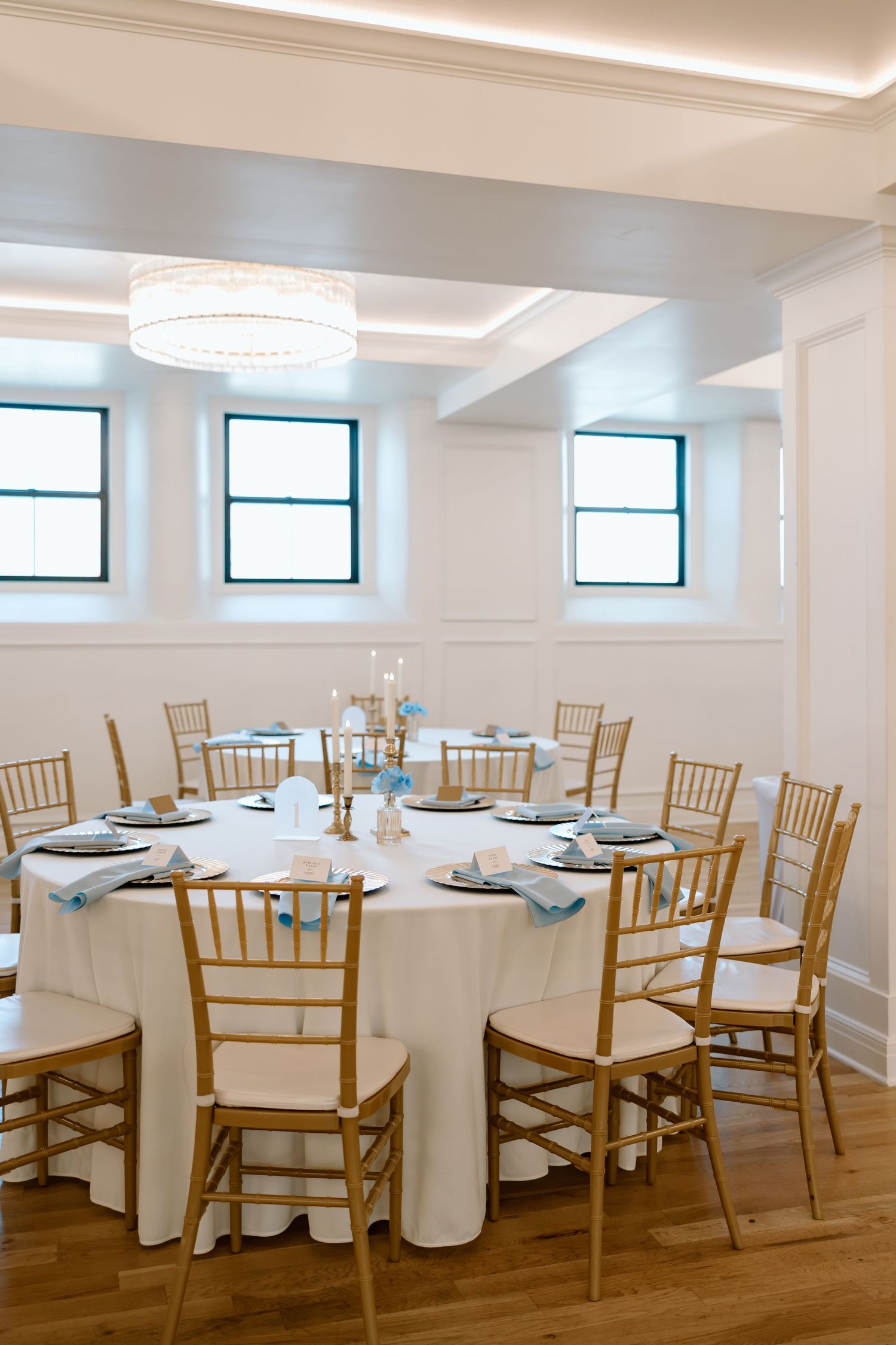 Elegant banquet room with round white-clothed tables, gold chiavari chairs, and a drum chandelier in a bright white venue.
