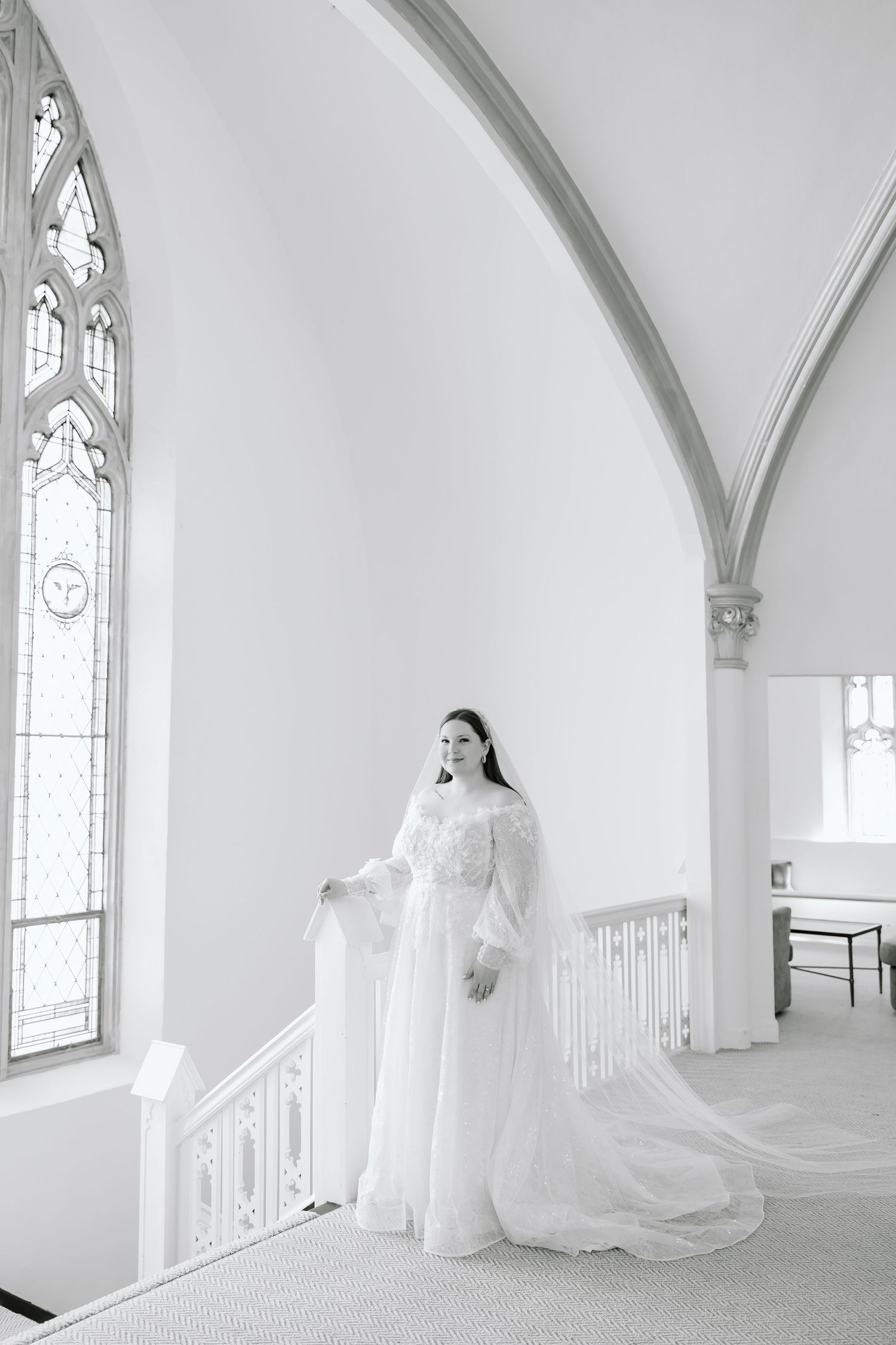 Bride in white gown and veil stands by staircase railing in elegant white church with Gothic arched windows.