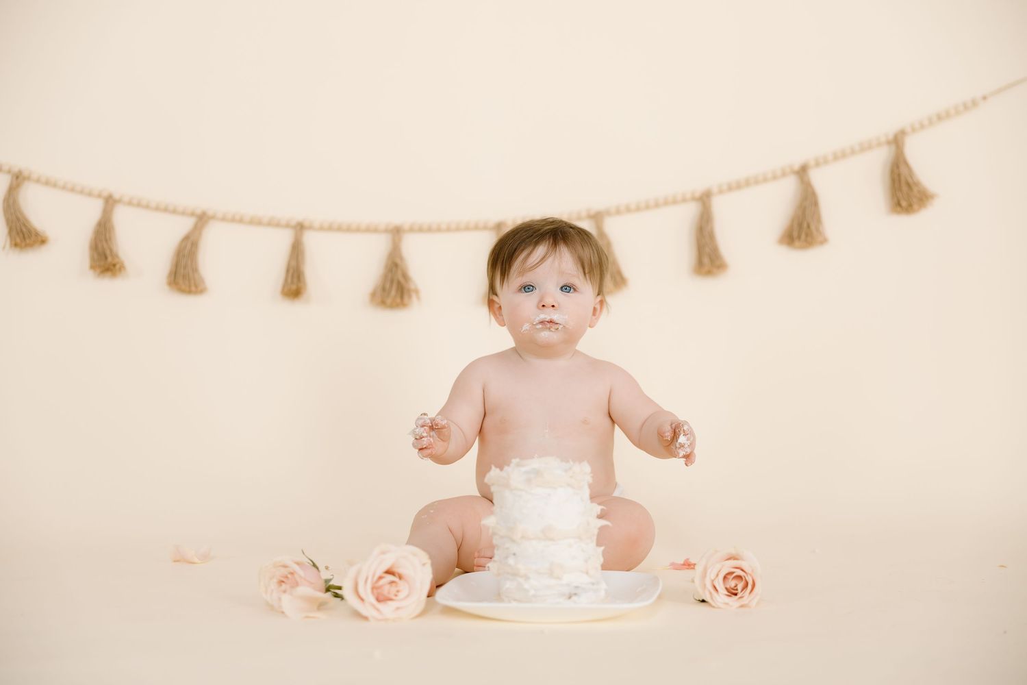 Baby seated with white cake and pink roses beneath decorative tassel garland on beige background.