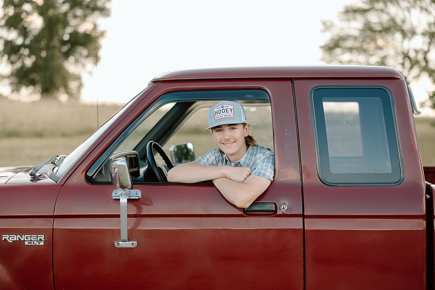 Someone wearing a baseball cap sits in the driver's seat of a maroon vintage Ford truck.