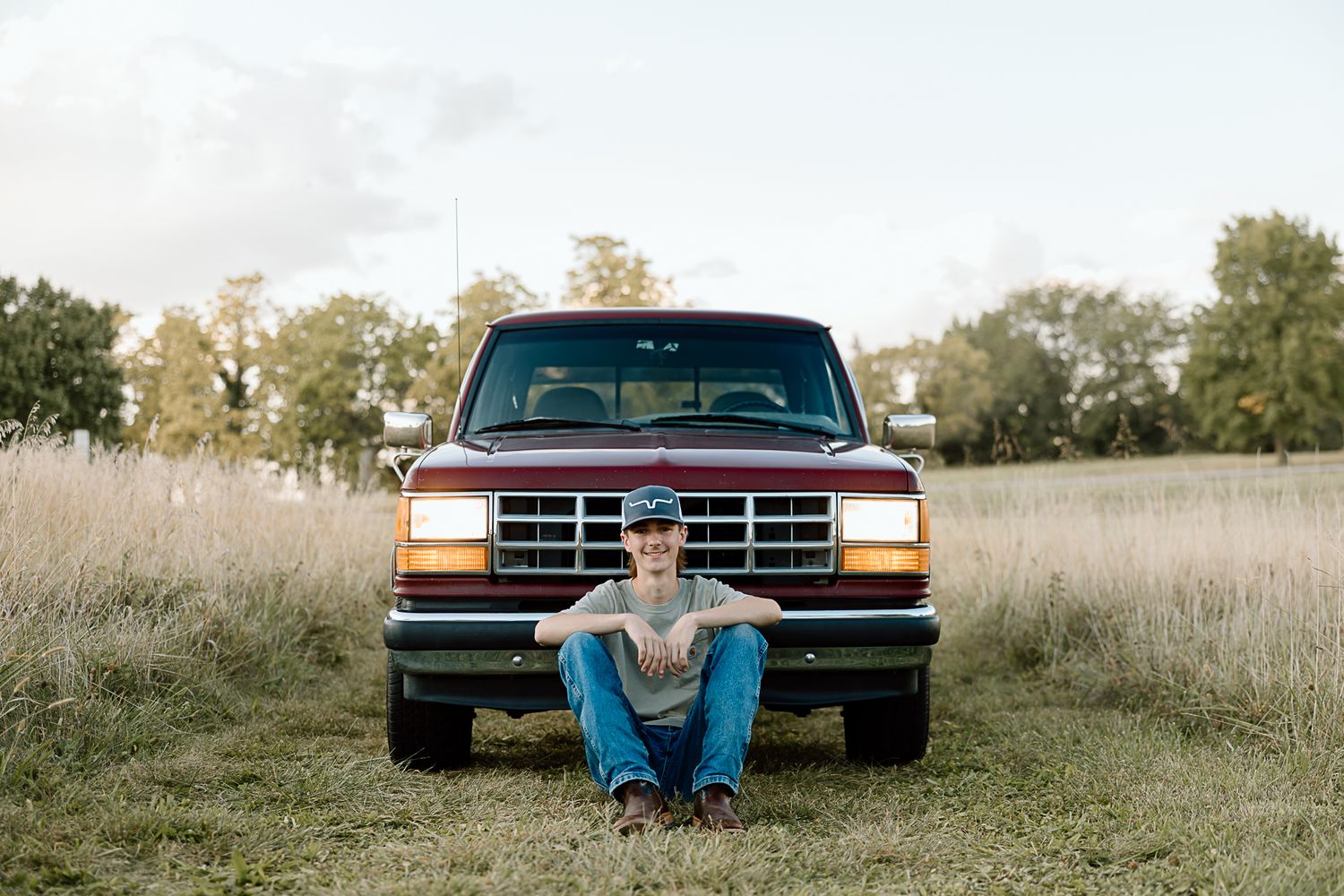 Someone sitting cross-legged in front of a red Ford pickup truck in a rural field setting.