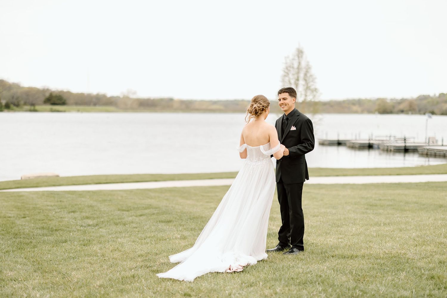 A wedding couple stands on a scenic lakeside lawn during their outdoor ceremony.