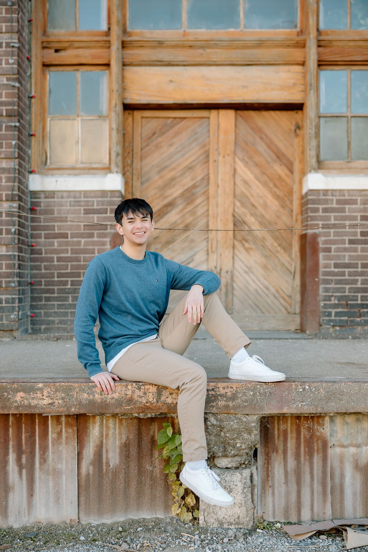 Casual outdoor portrait with person in blue sweater sitting on weathered concrete ledge near wooden doorway.