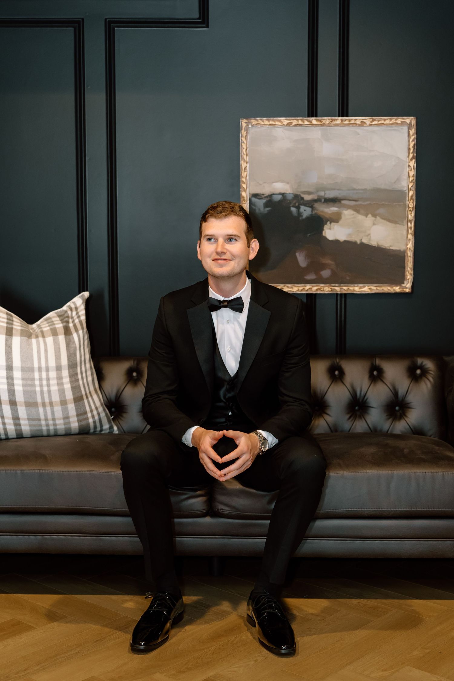 A young man in a black tuxedo sits on a tufted leather sofa against a dark wall with a landscape painting.