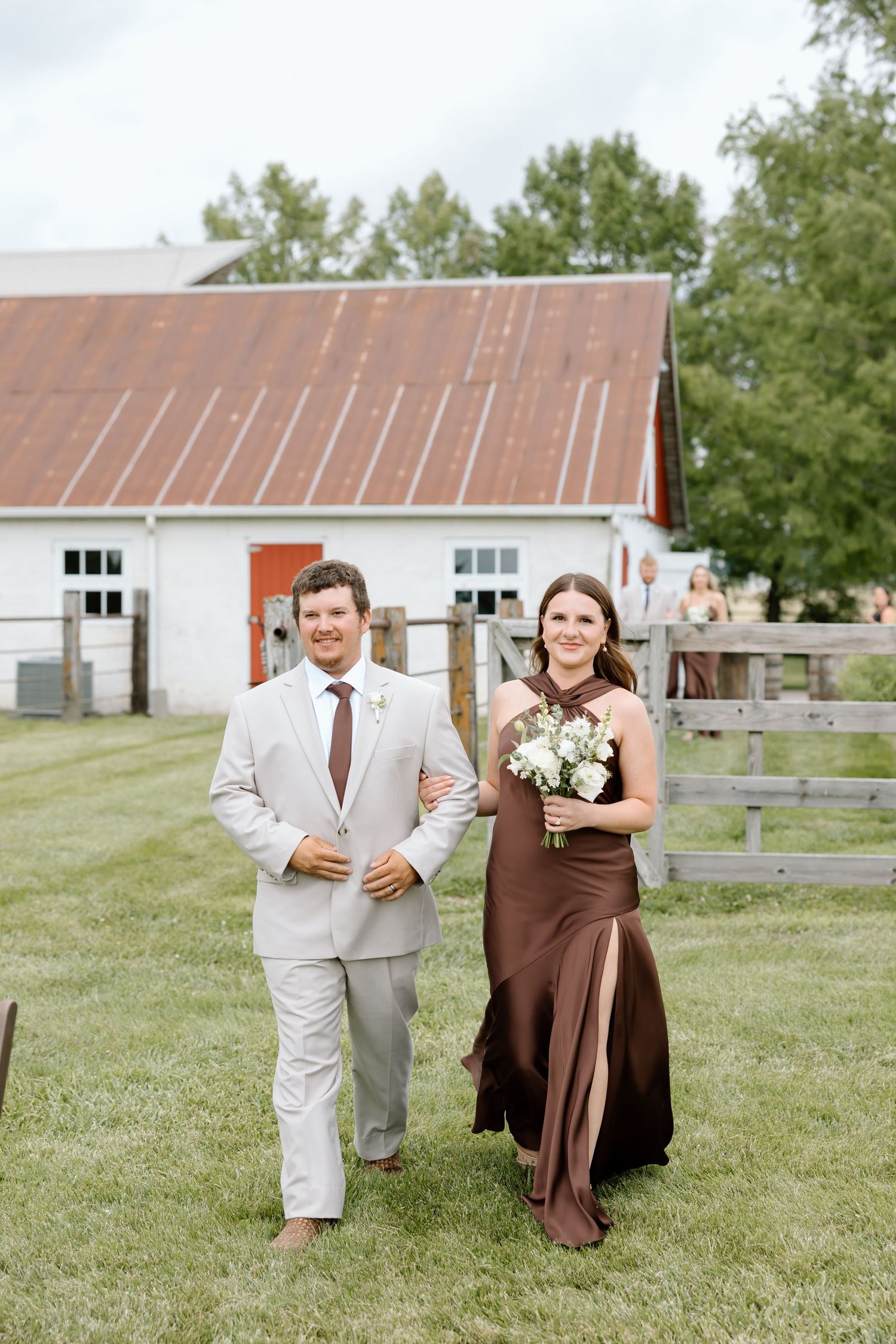 A groomsman in a tan suit escorts a bridesmaid in a brown dress at an outdoor barn wedding ceremony.